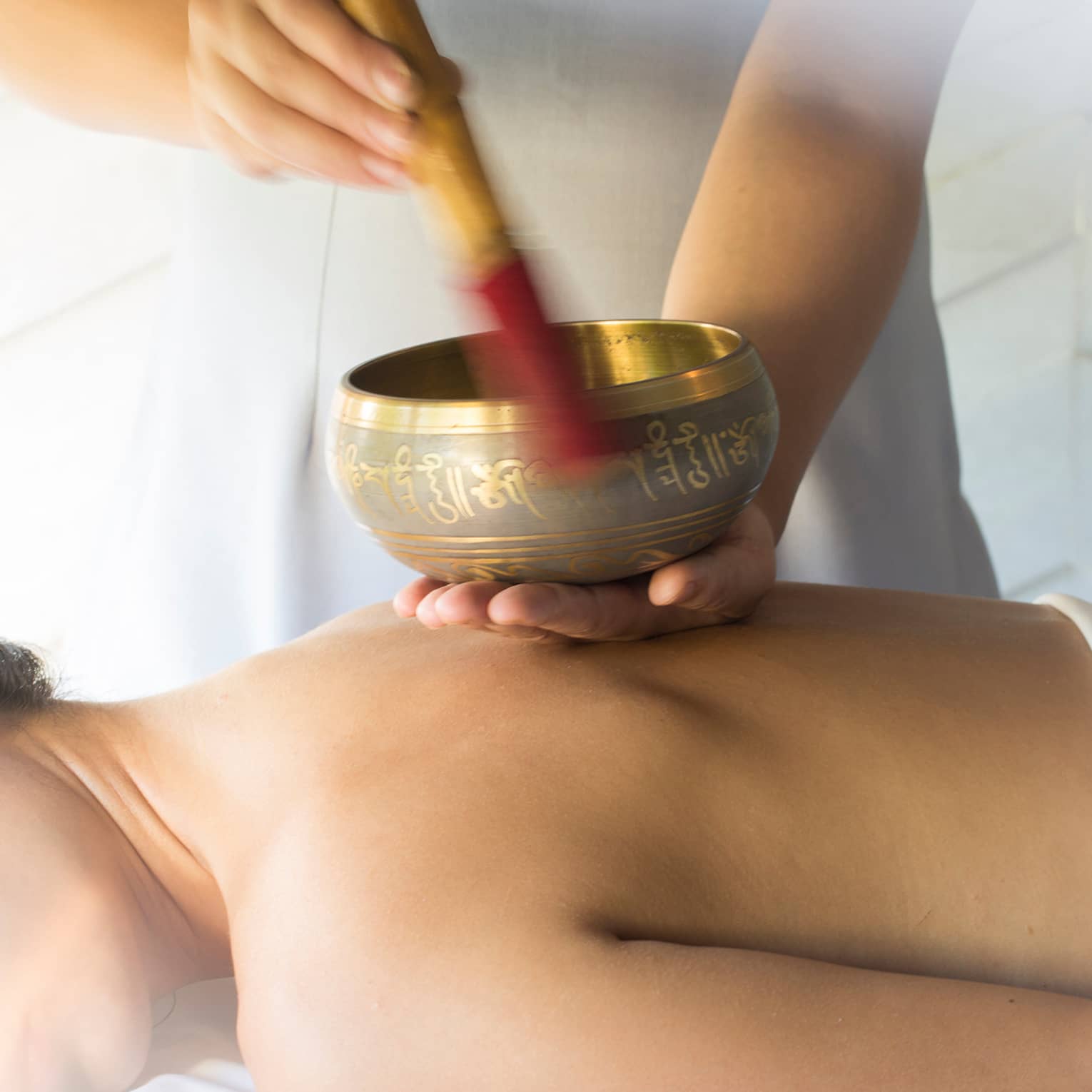 Hands play brass singing bowl over bare back of woman lying on massage table