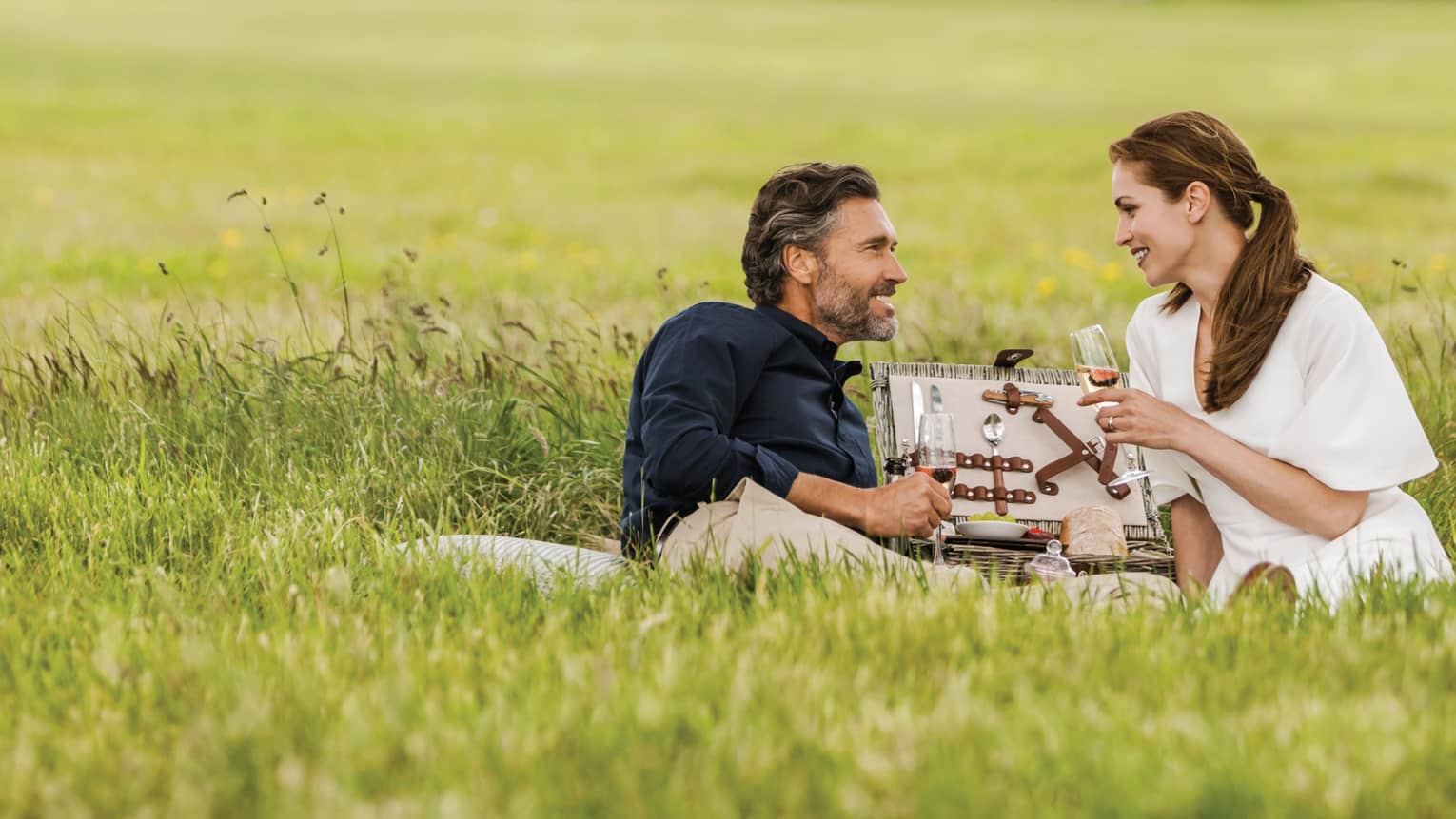 Couple with wine glasses lies in tall green grass with open picnic basket, hotel manor in background