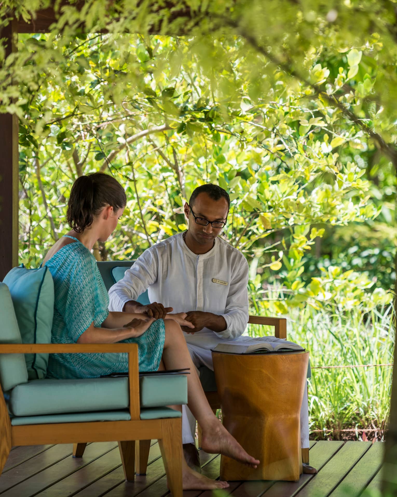 Spa staff places hand on woman's wrist by open book on plush blue chairs on patio pavilion