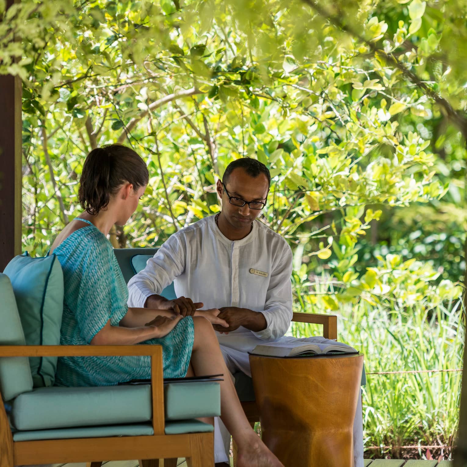 Spa staff places hand on woman's wrist by open book on plush blue chairs on patio pavilion