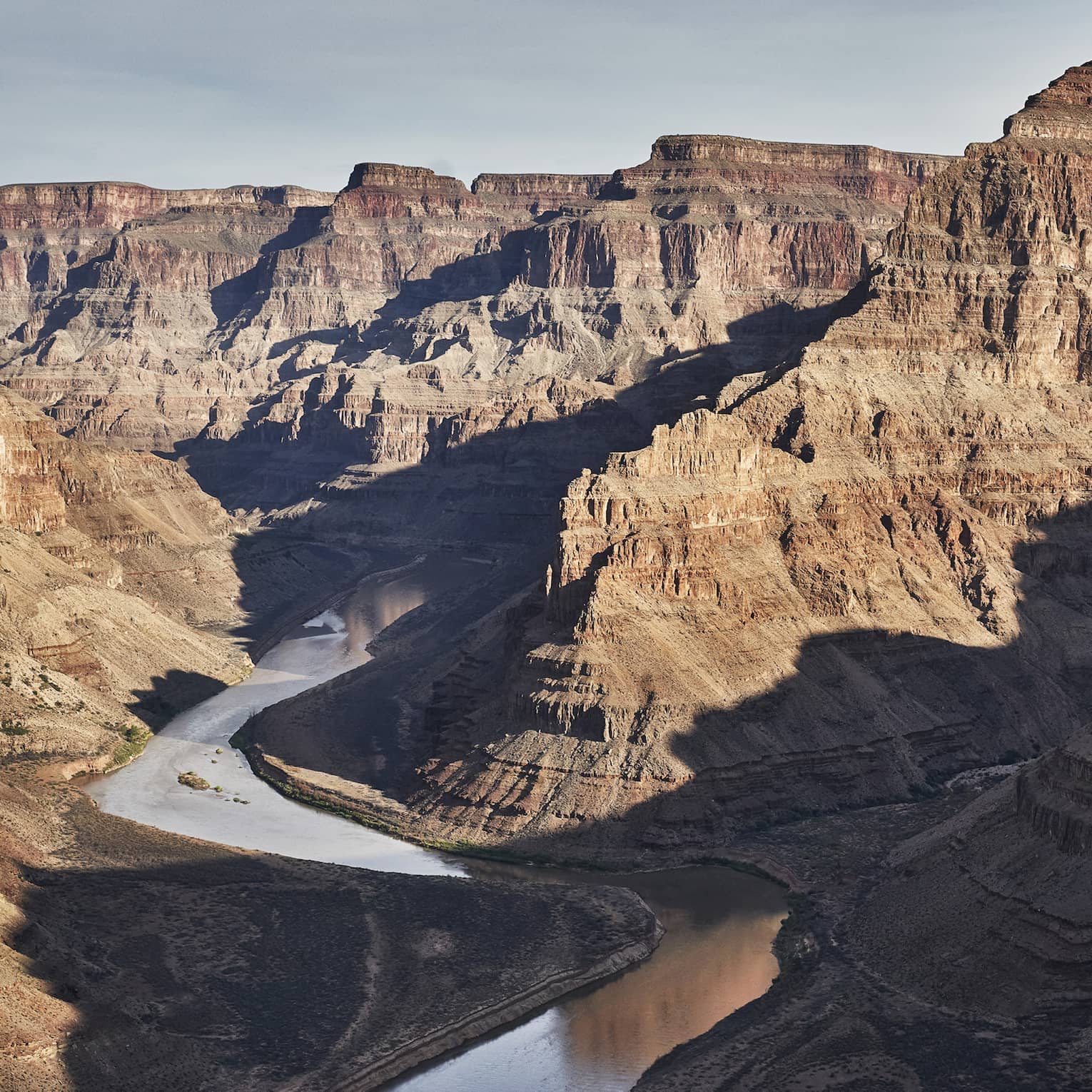 A silvery river meanders its way along the bottom of a wide, deep canyon with flat-topped ridges of rock towering upwards.