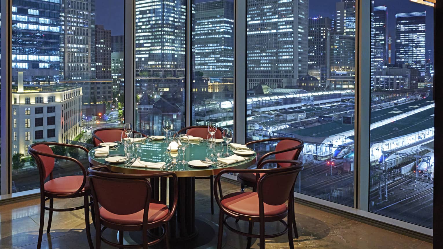 Luxury boutique hotel dining setup with a round table elegantly set for guests, offering a panoramic city view at night through tall glass windows
