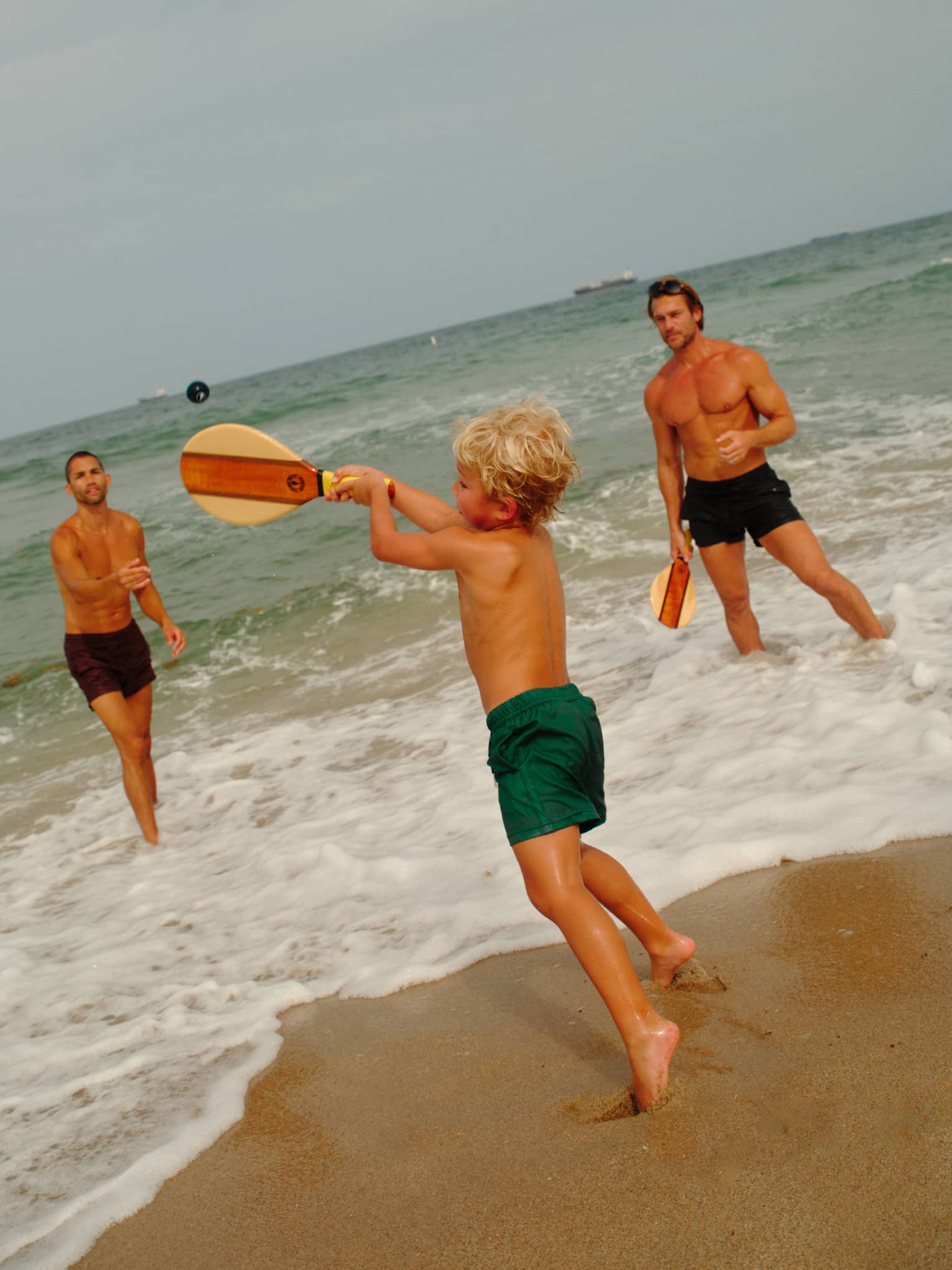 A young shild plays paddle ball in the surf with two adults
