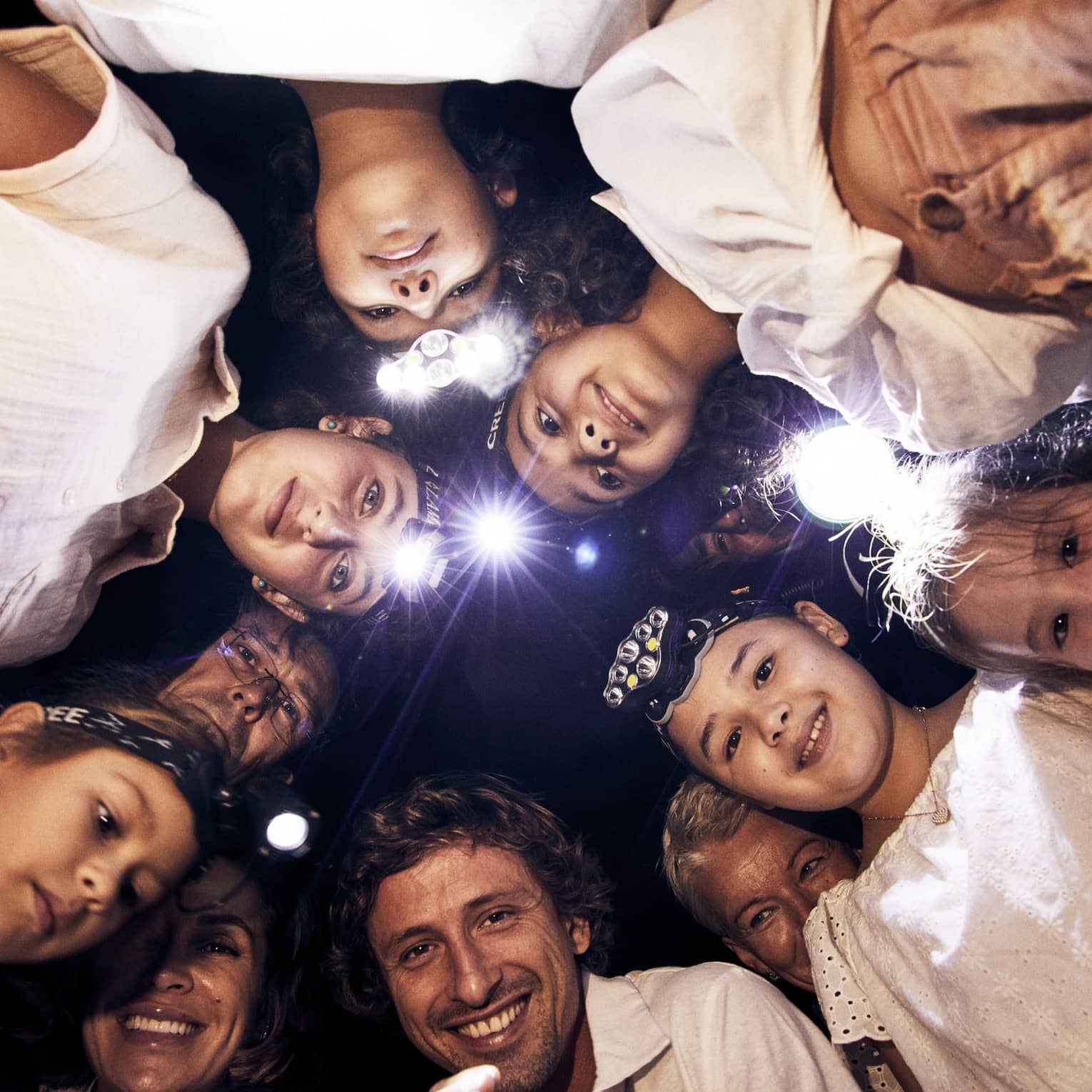 A group of children and adults, some wearing headlamps, stand in a circle and smile downwards into the camera. 