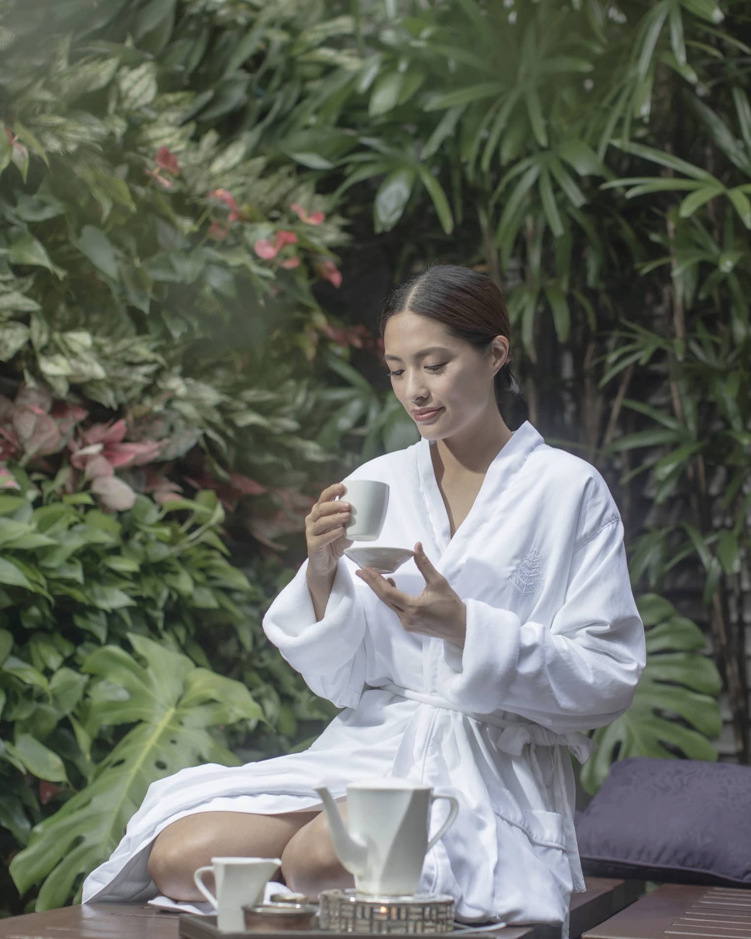 Woman in white spa robe kneeling and sipping tea among outdoor foliage