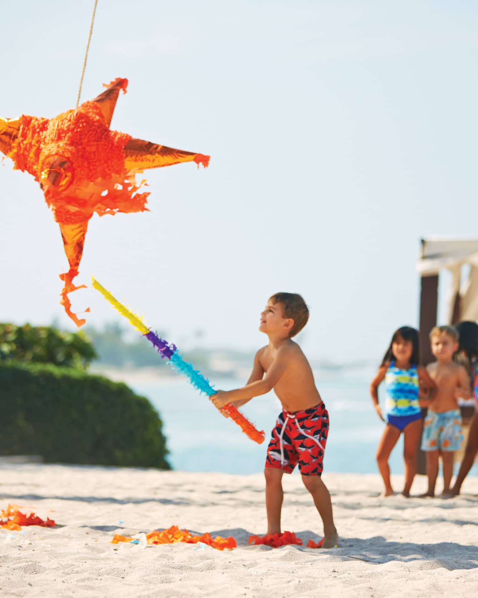 On a beach, a child swings a bat wrapped in vibrant crepe paper at a big orange star piñata; other children watch excitedly. 