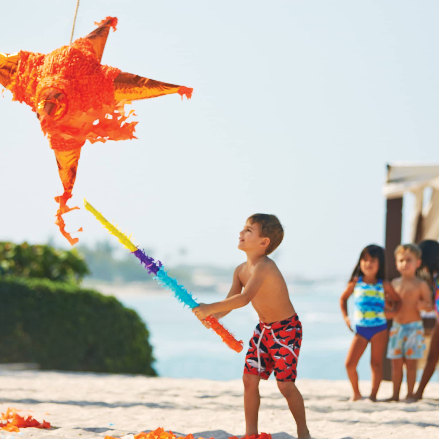 On a beach, a child swings a bat wrapped in vibrant crepe paper at a big orange star piñata; other children watch excitedly. 
