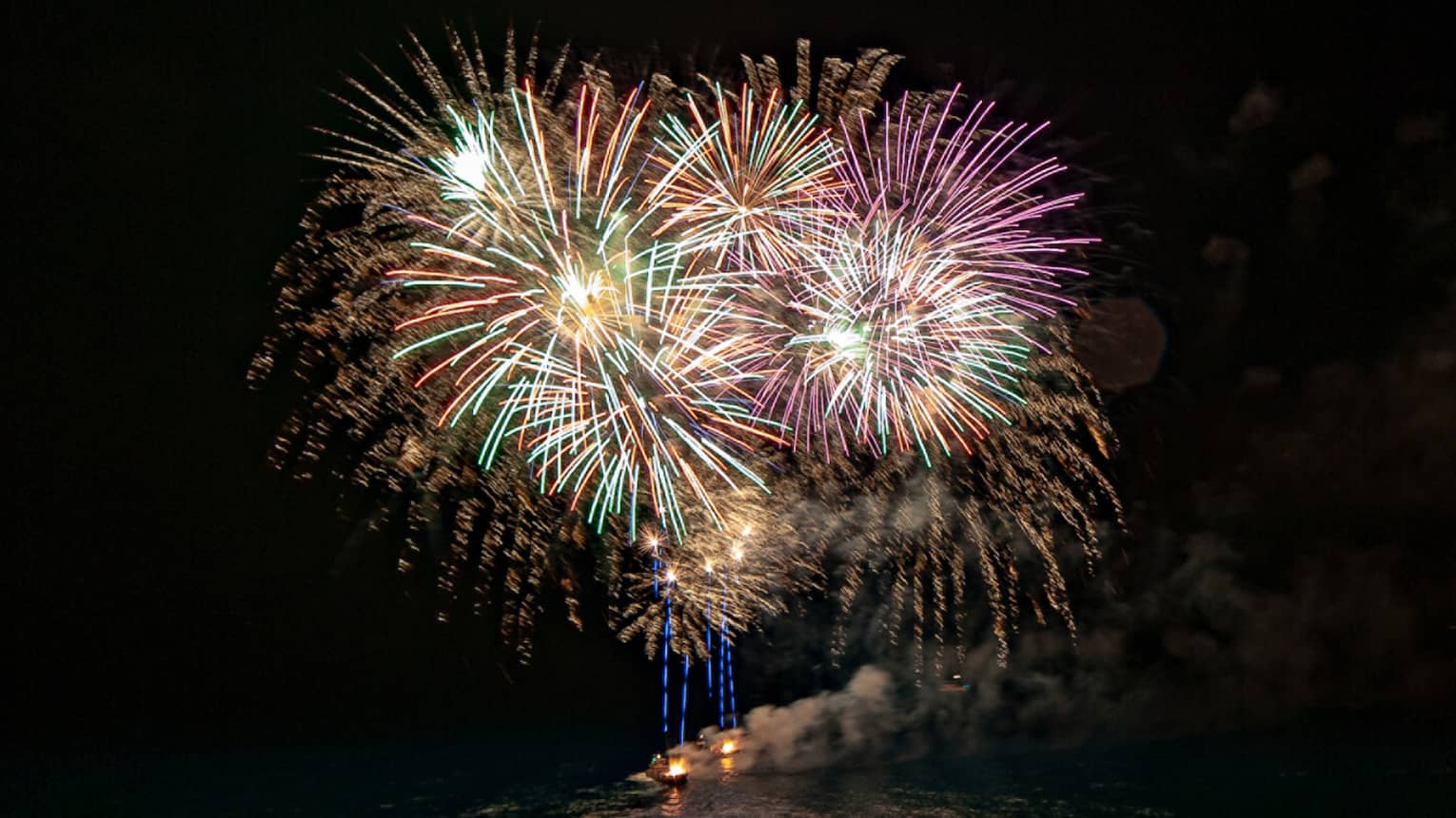 Fireworks shot from boats explode in a chrysanthemum effect of yellows, greens, oranges and pinks against a pitch-black sky.