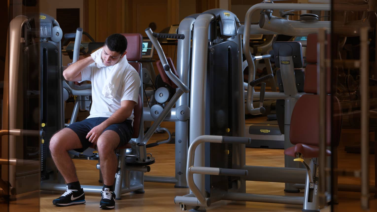 Man sits on weights machine, white towel around neck, past orange glass doors in fitness centre