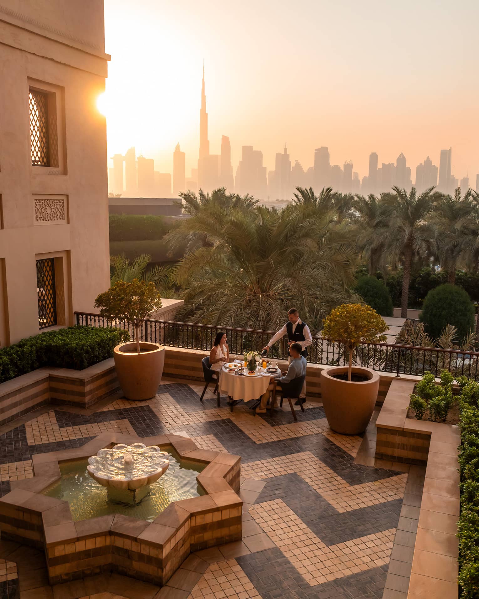 Table set for two on an open-air rooftop terrace with a chevron tiled floor and the sun rising over a city skyline in the near distance