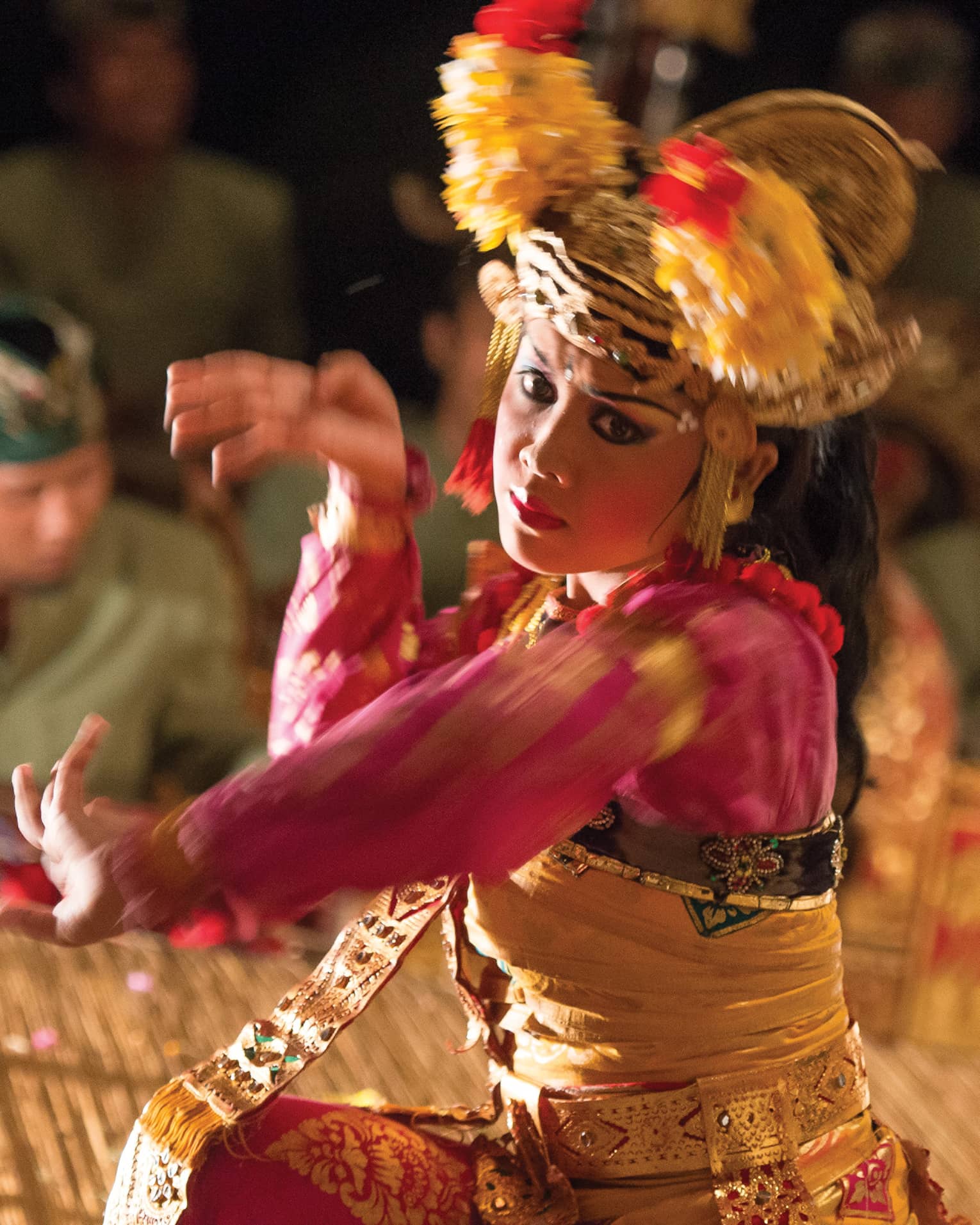 Close-up of a Balinese dancer in a golden headdress and bright clothing; in the background musicians play the gamelan. 