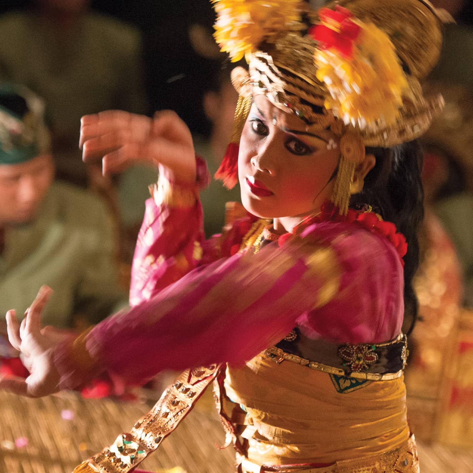 Close-up of a Balinese dancer in a golden headdress and bright clothing; in the background musicians play the gamelan. 