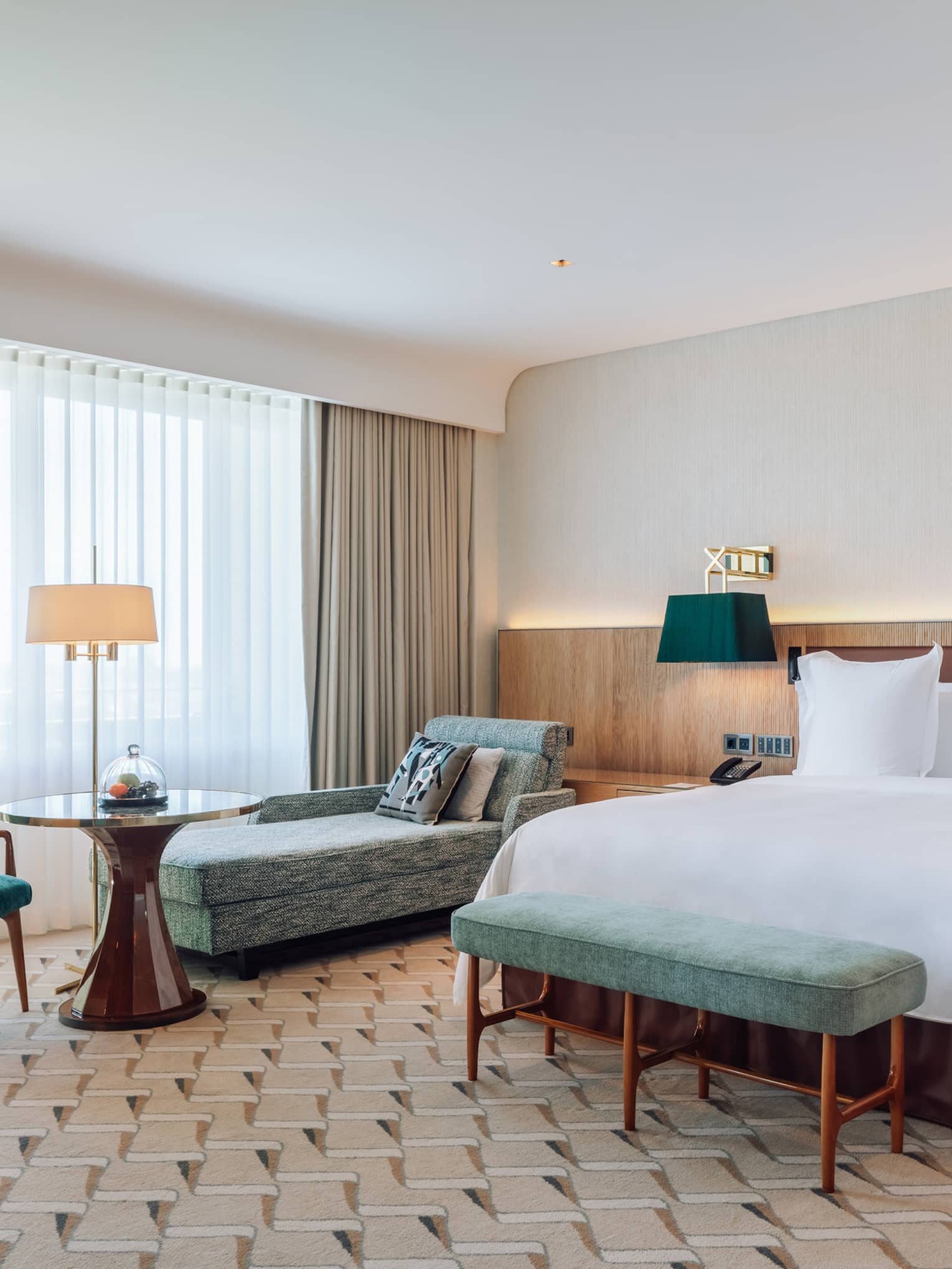 Modern hotel room with a large bed, chaise lounge, dining table, green chairs and floor-to-ceiling windows.