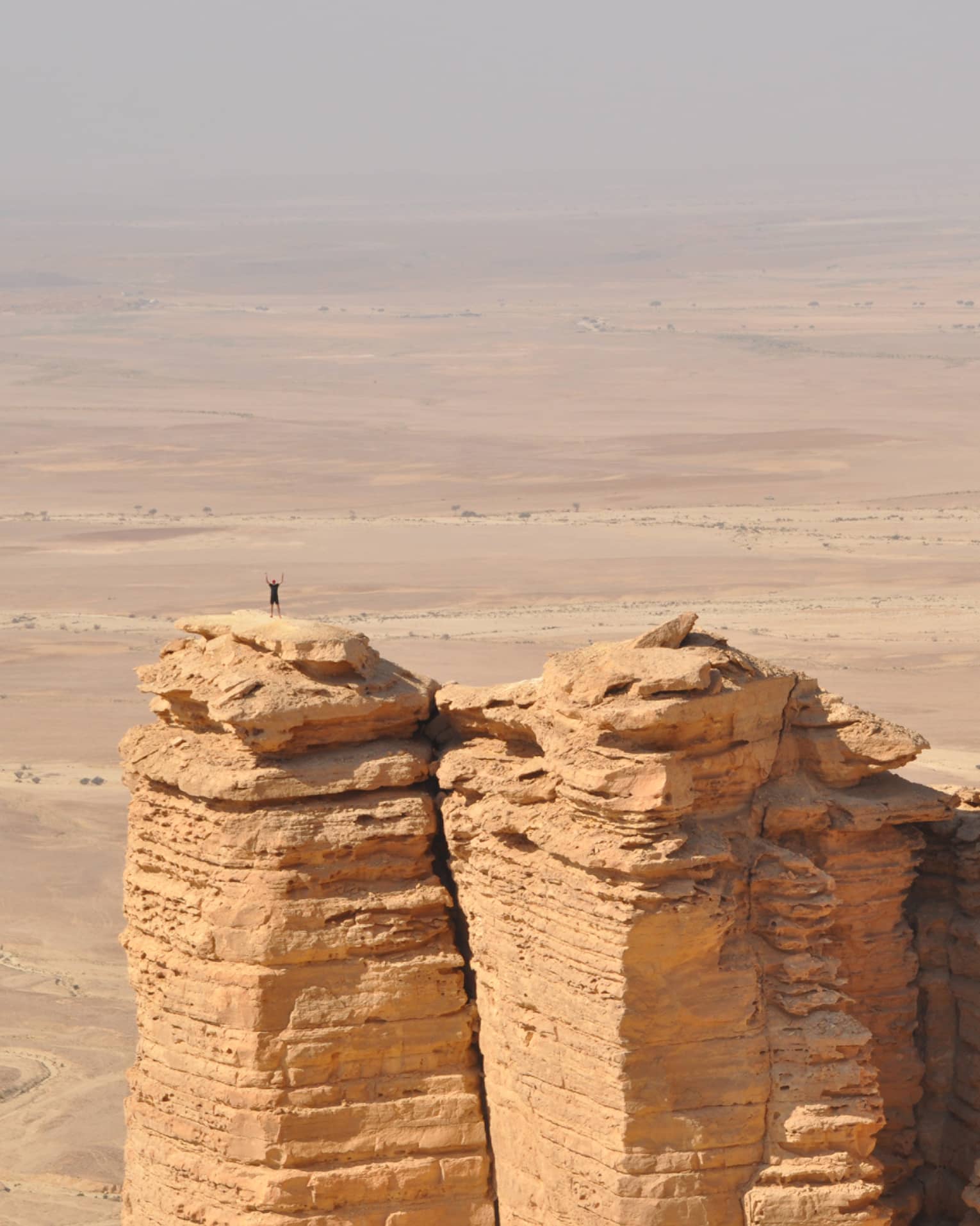 An aerial view of a giant golden cliff surrounded by a dry desert and a small silhouette of a person standing on top of it.