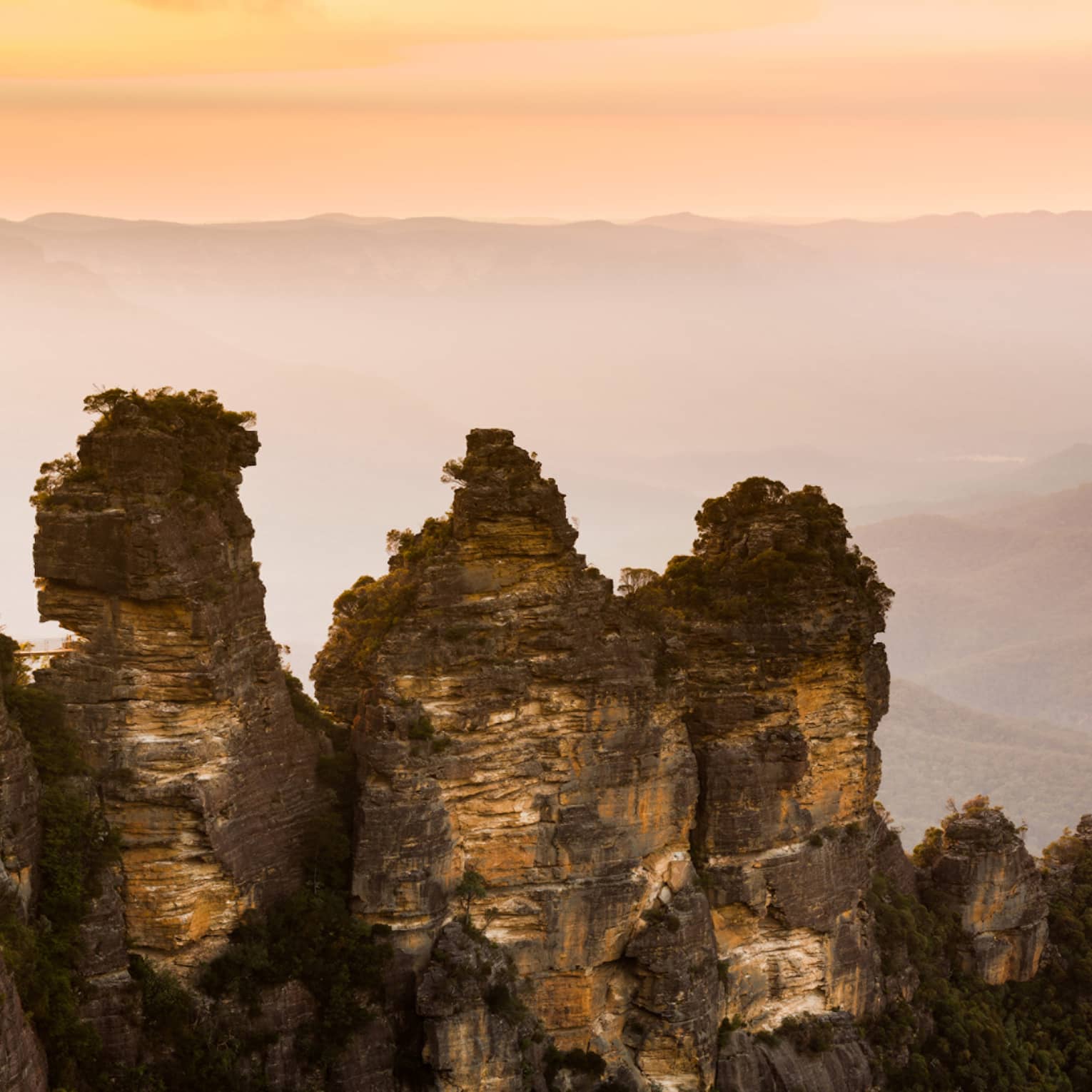 Dramatic sunset view looking out across misty mountains, a wall of craggy, sparsely treed rocks rising up in the foreground.