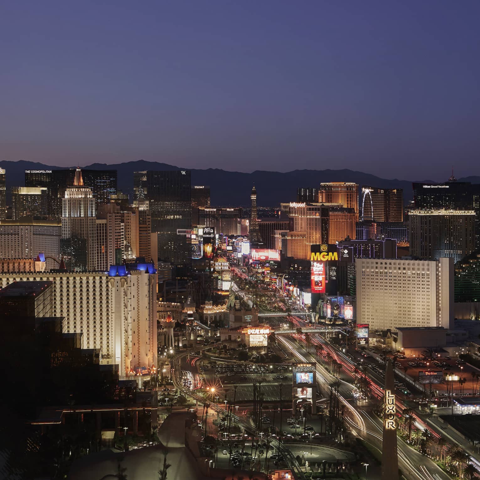 Aerial view of the Las Vegas Strip at night with lit-up buildings and neon signs, low mountains beyond under a dark blue sky.