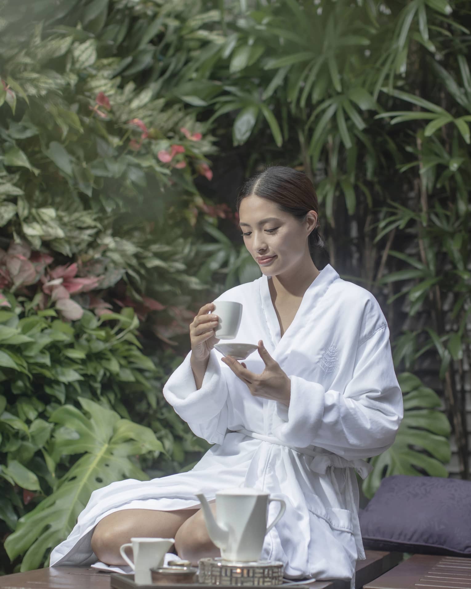 Woman in white spa robe kneeling and sipping tea among outdoor foliage