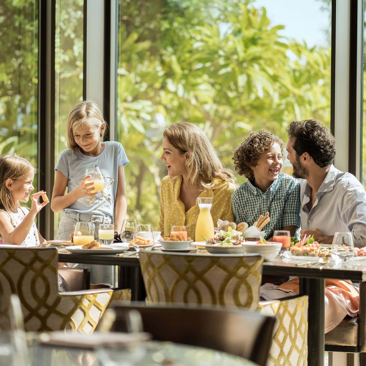 A family of five eating breakfast on the same side of the table, with trees in the background.