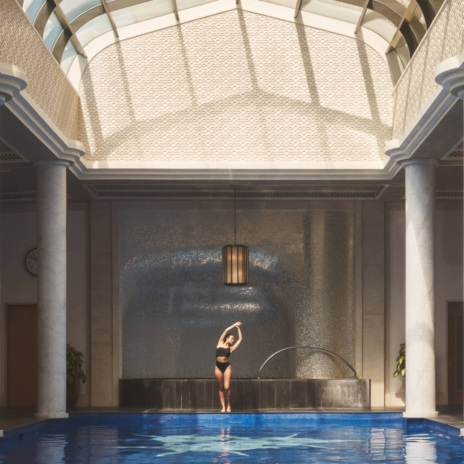 Person in swimsuit standing and stretching with arms overhead beside indoor pool flanked with white pillars under vaulted ceiling