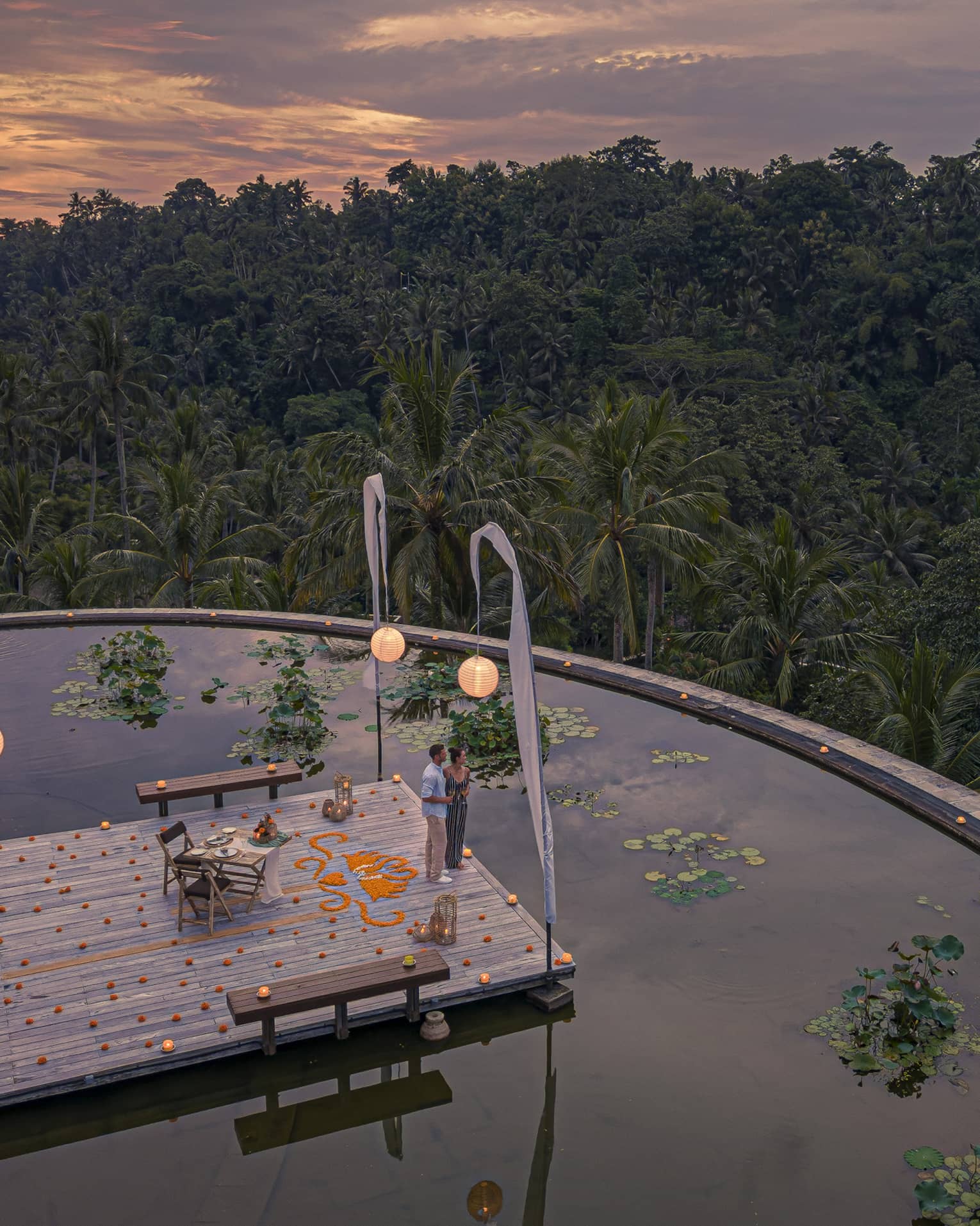 Couple stands on dock at the infinity edge lotus pond overlooking dense forest at dusk