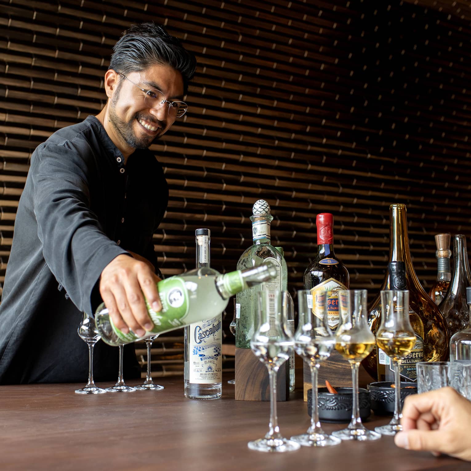 A bartender pours a clear liquor into grappa glasses, two others filled with an amber liquor, aside various other bottles.