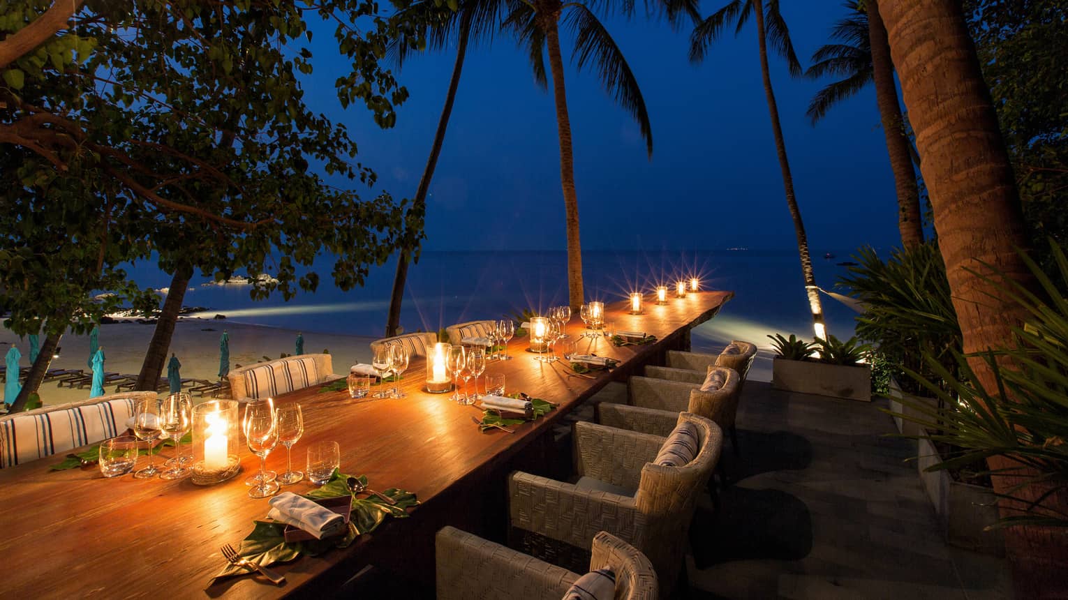 Long wood outdoor dining table with glowing candles under palm trees at night