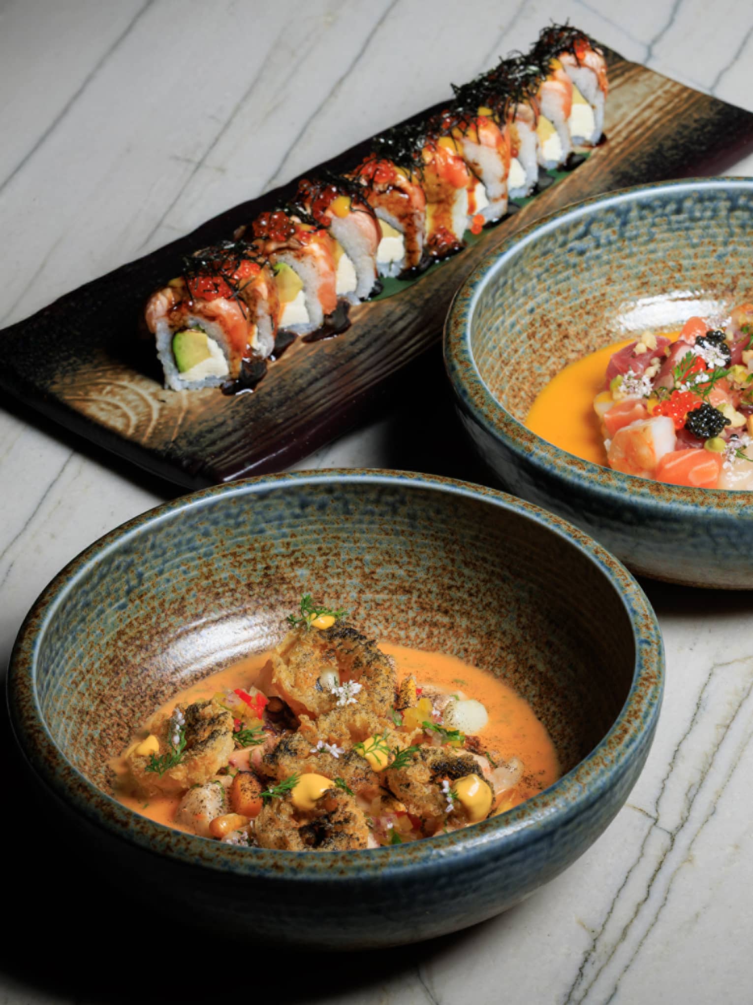 Two stoneware bowls filled with food sit next to a rectangular platter of sushi on a white wood-grain table