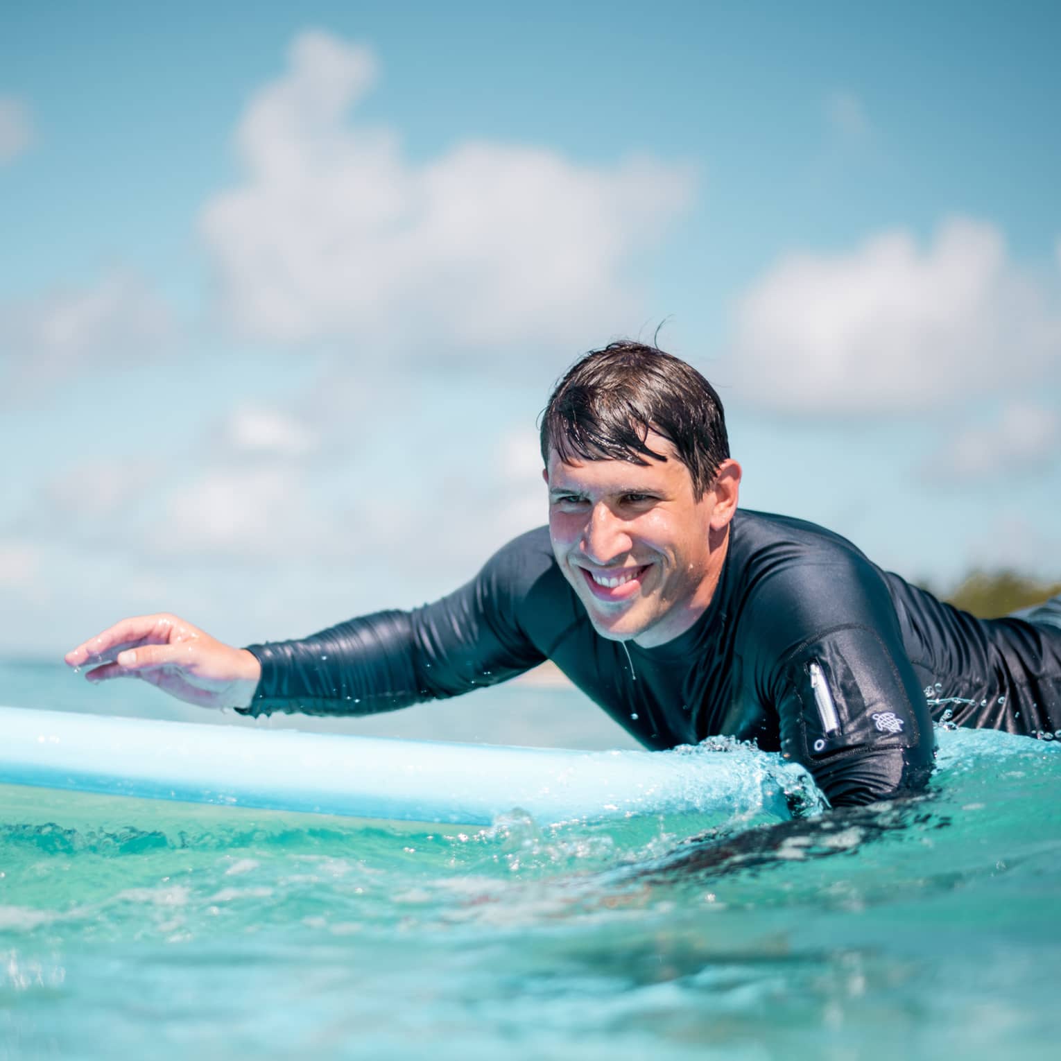 Man in rash guard smiling while paddling on blue surfboard in blue-green water