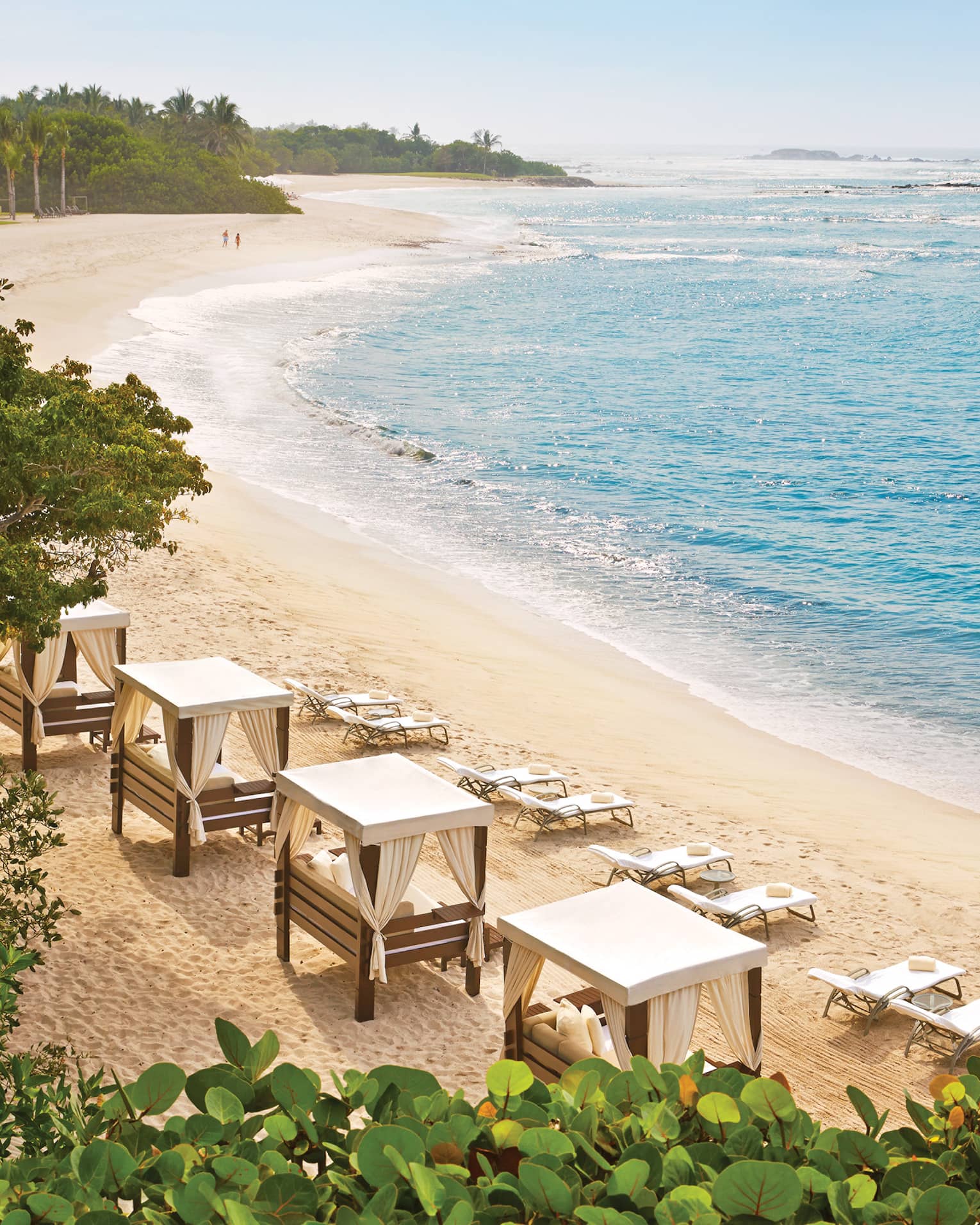 Amid lush vegetation, azure waves roll onto a curved beach lined with chairs and daybeds. A couple strolls in the distance.