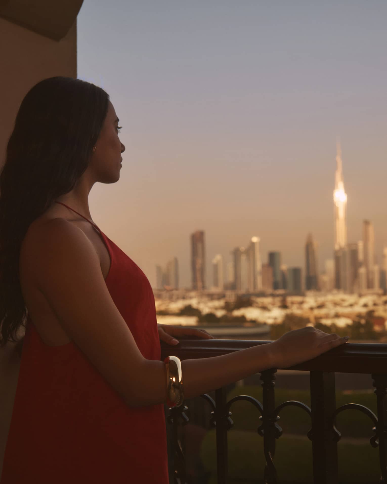 Person with long brown hair standing on balcony with hand resting on railing, looking out to a city at dusk