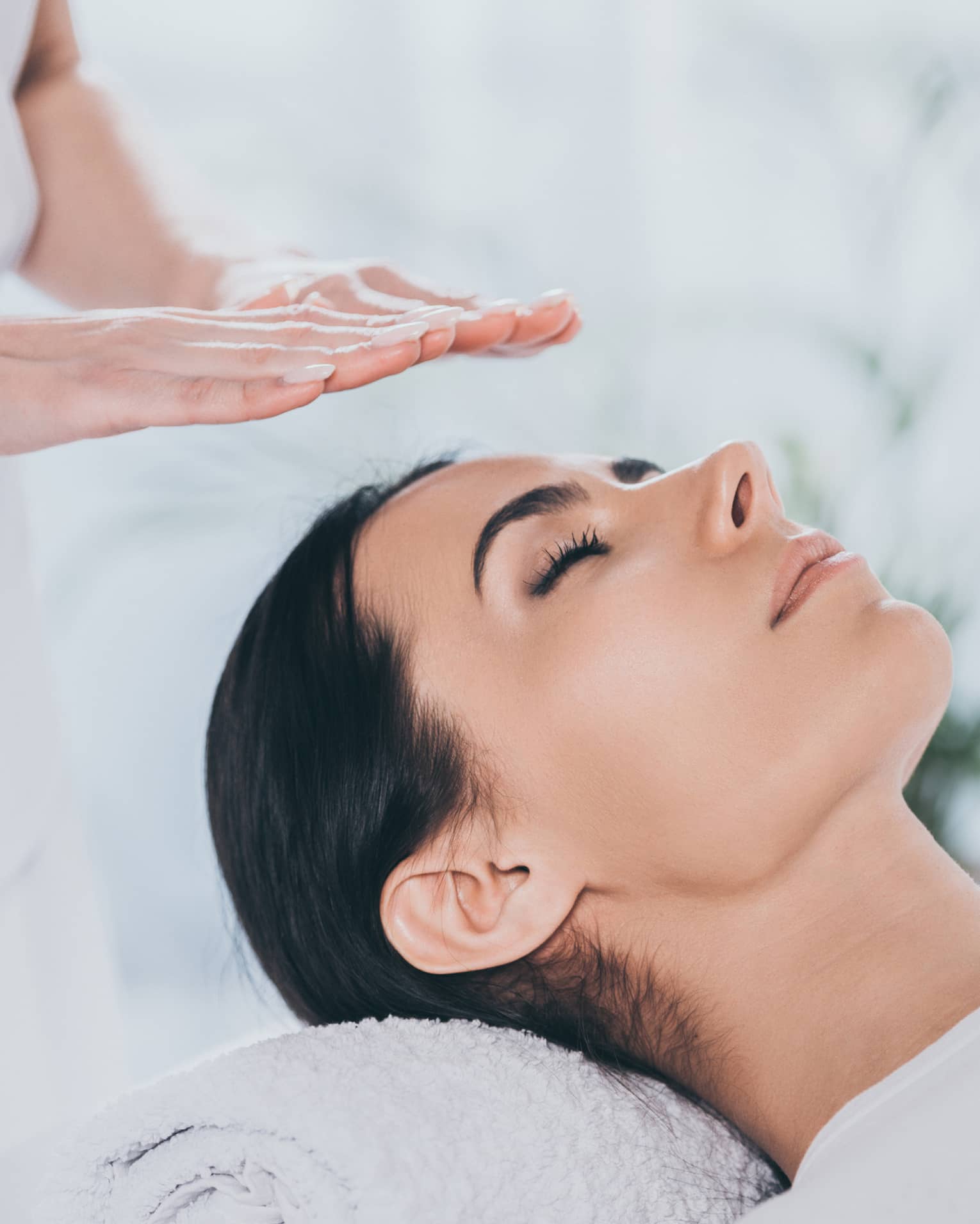 Woman with eyes closed lays on rolled up towel on spa table under therapists' hovering hands