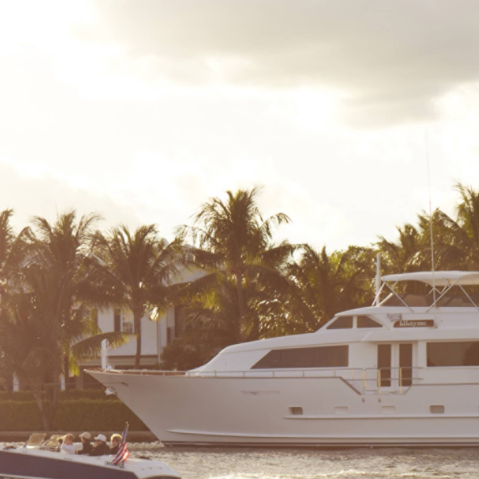 A large white yacht cruises alongside a smaller boat in a serene waterway, with palm trees and luxurious homes in the background under a partly cloudy sky.