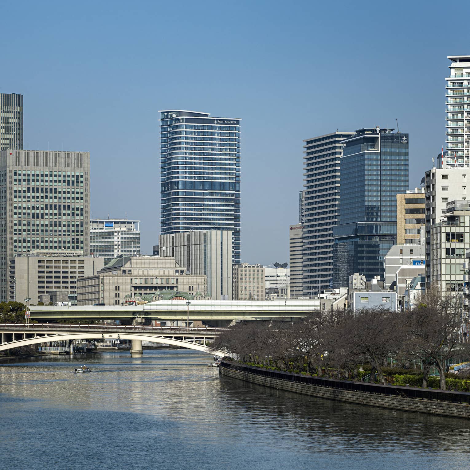 Under a clear blue sky, Osaka's modern high-rises loom above two bridges spanning a river lined with dormant cherry blossoms.