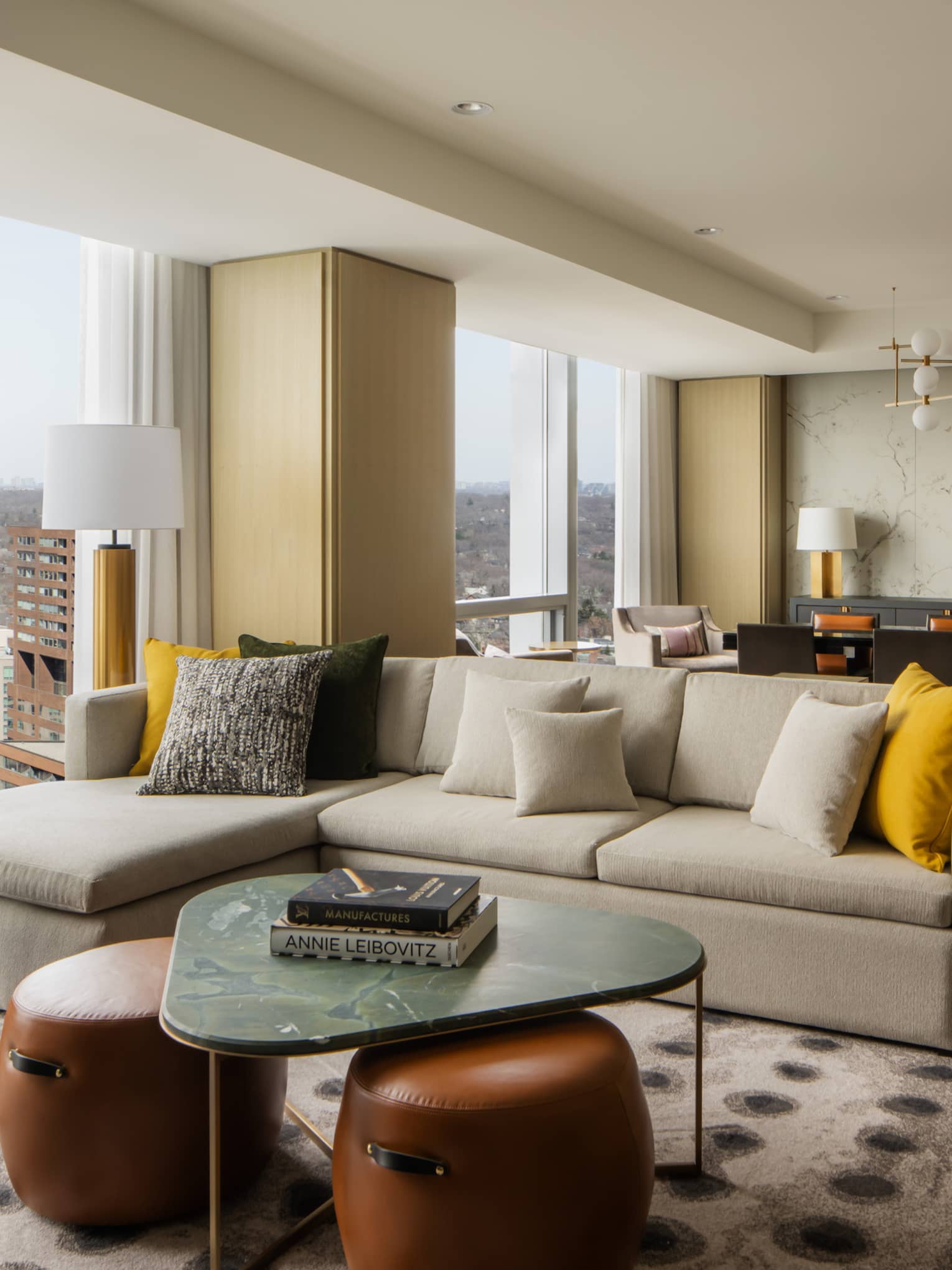 Spacious suite living area with a large sectional sofa, yellow and gray pillows, marble coffee table, and a dining area with elegant décor and city views