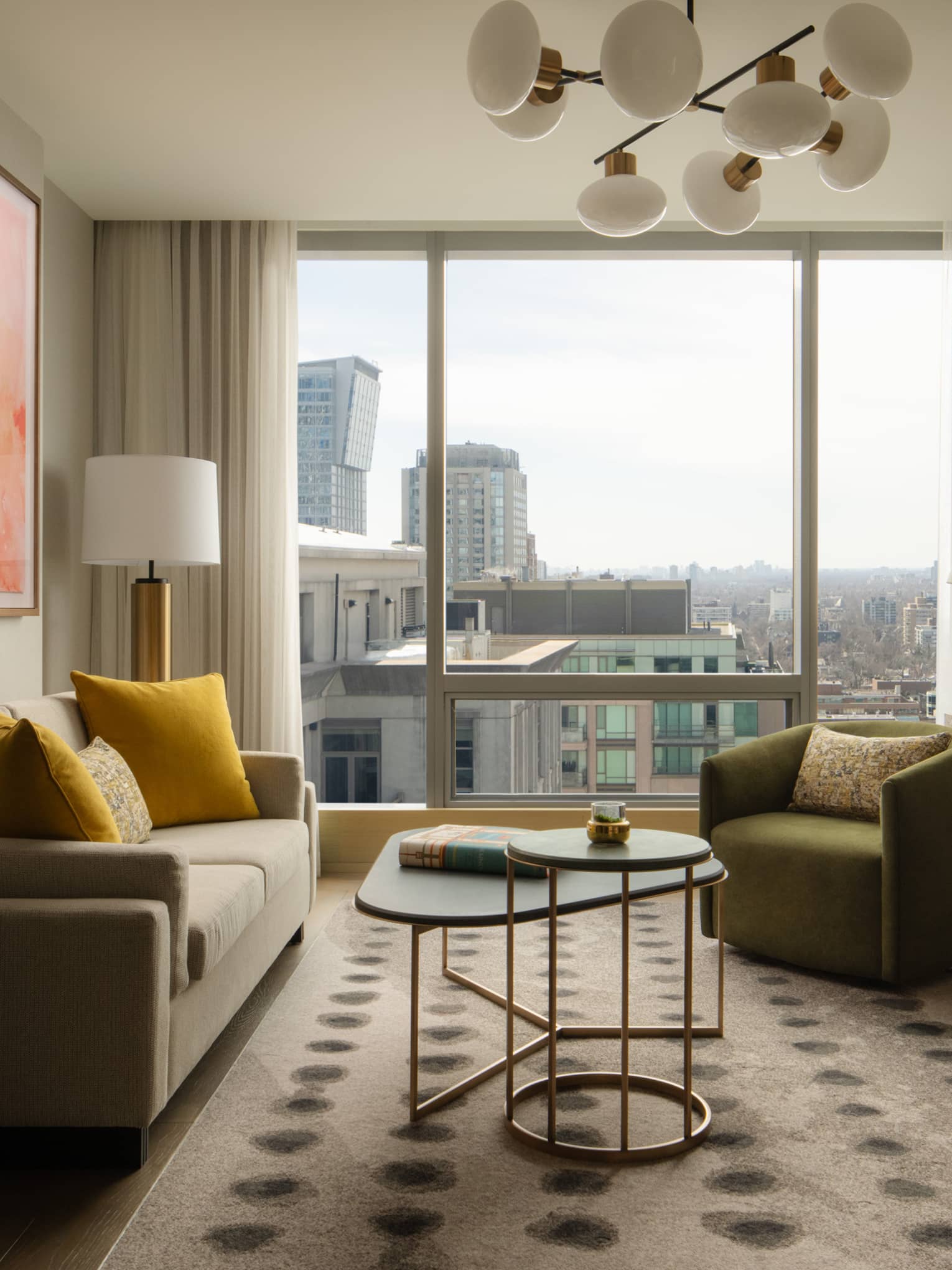 Hotel suite living room with a grey sofa, green armchair, coffee tables, console table with TV, abstract artwork and large windows
