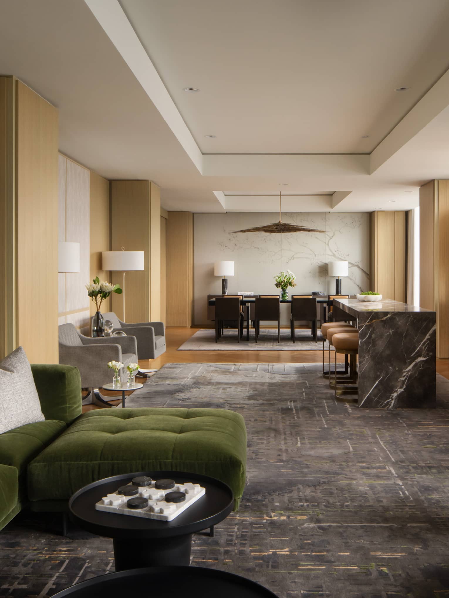 Luxury hotel suite lounge area with a green velvet sectional, marble-top bar, modern dining table, and open shelving with decorative accents