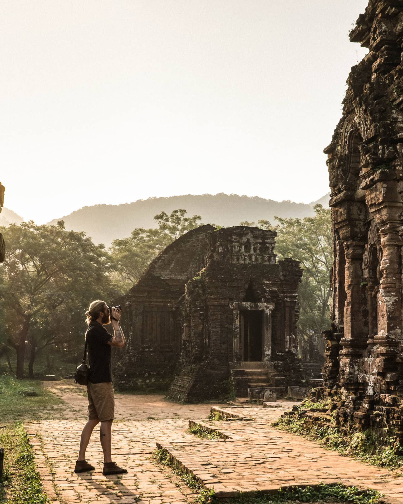 A tourist photographing ancient stone ruins, surrounded by lush greenery and misty hills in the background.