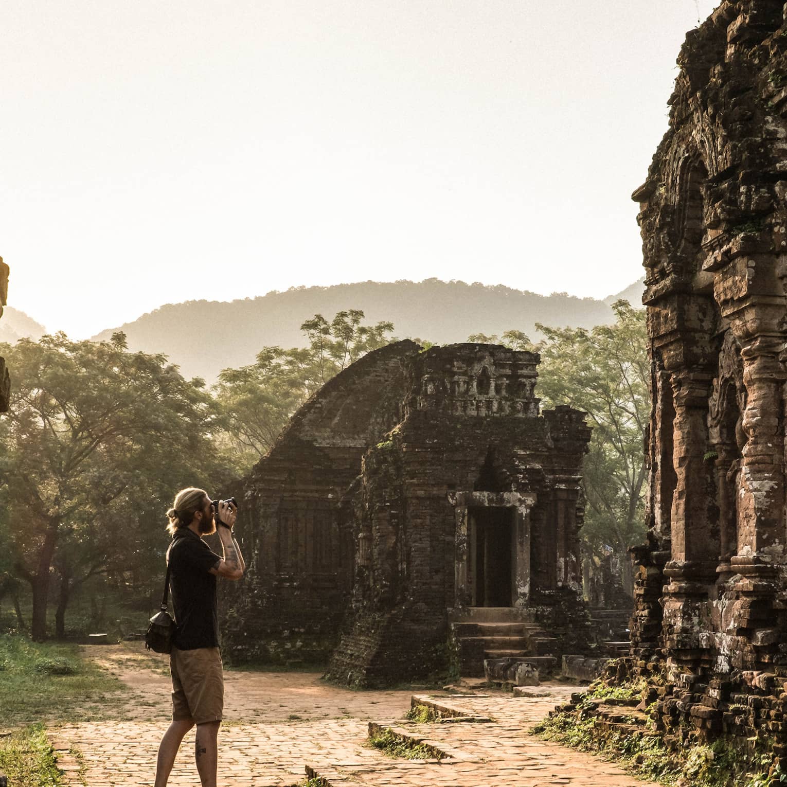 A tourist photographing ancient stone ruins, surrounded by lush greenery and misty hills in the background.