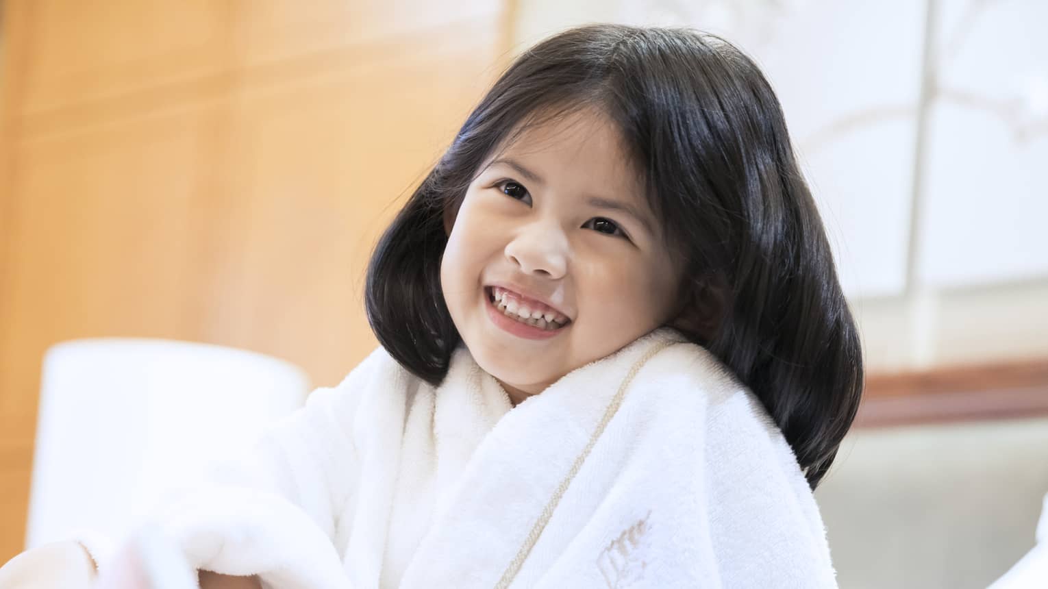 Smiling little girl wearing white bath robe and pink slippers sits on bed