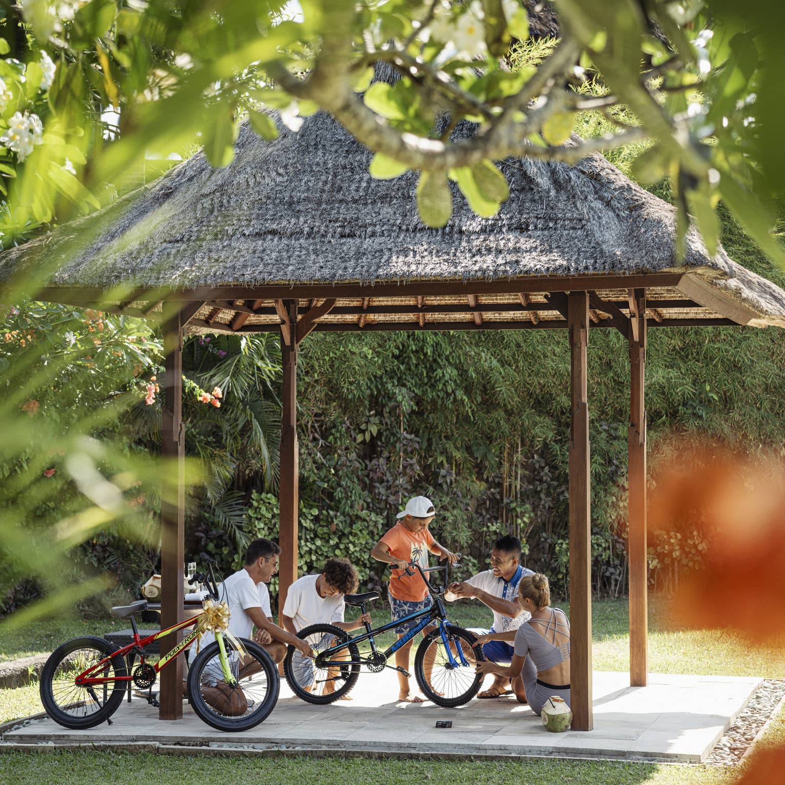 View through foliage of four adults and a child working on a bicycle under a thatched hut surrounded by lush greenery.