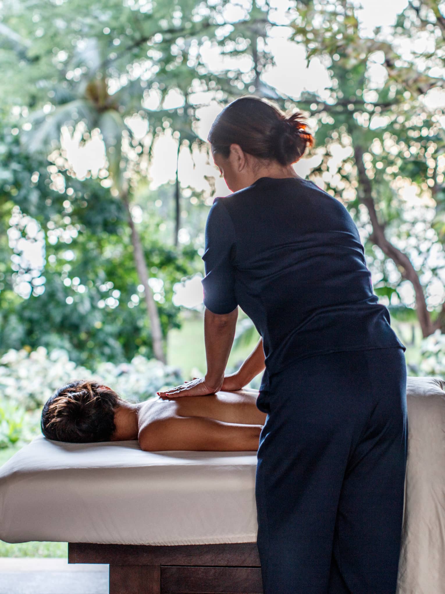 Spa staff massages woman's bare shoulders as she lies under sheet on massage table