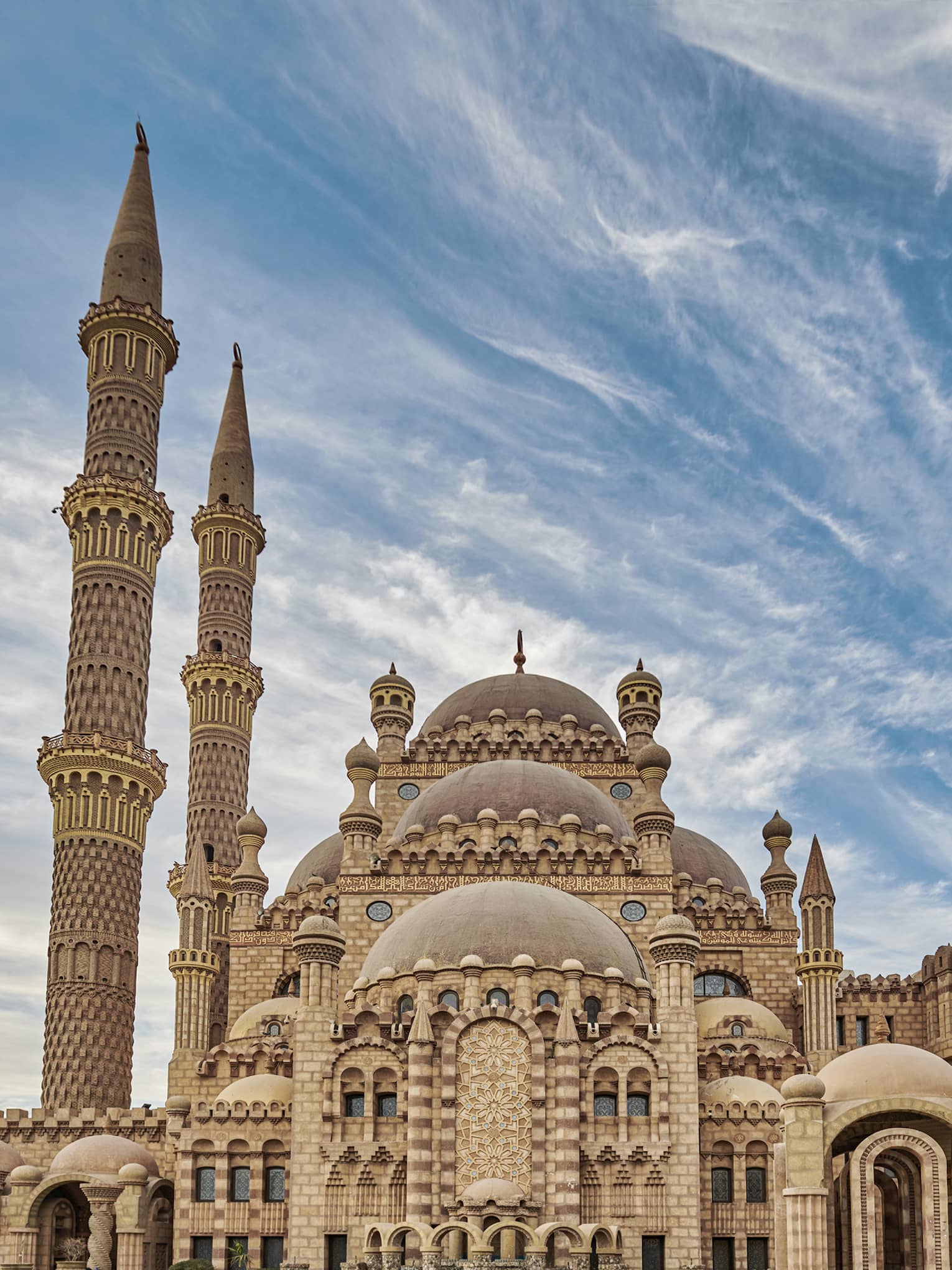 Full view of a sandstone mosque. It has many copper domes, two minarets on the left, and elaborate geometric carvings.