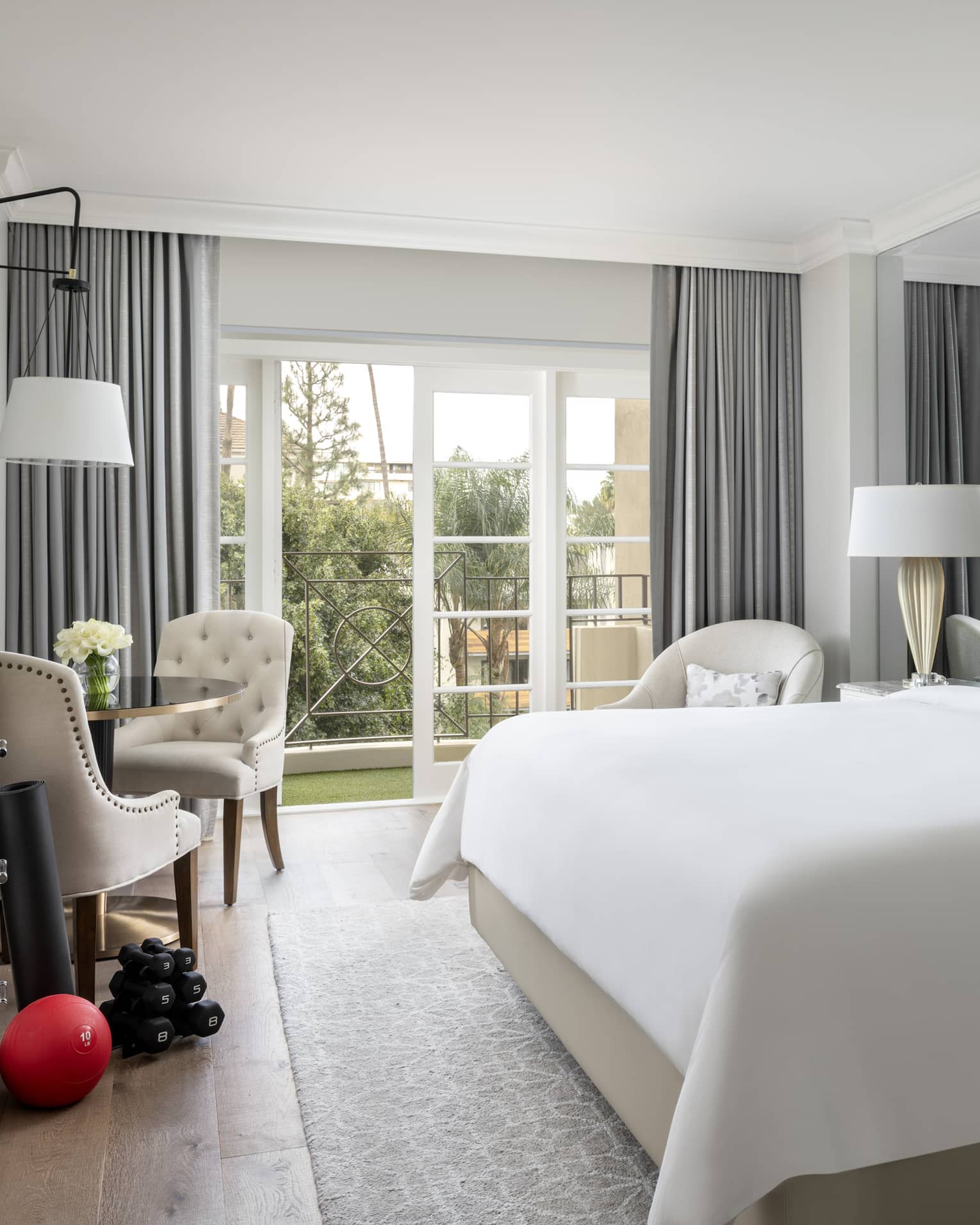 A hotel room with large open window, bed with white sheets, dresser, two chairs, an exercise ball and weights.