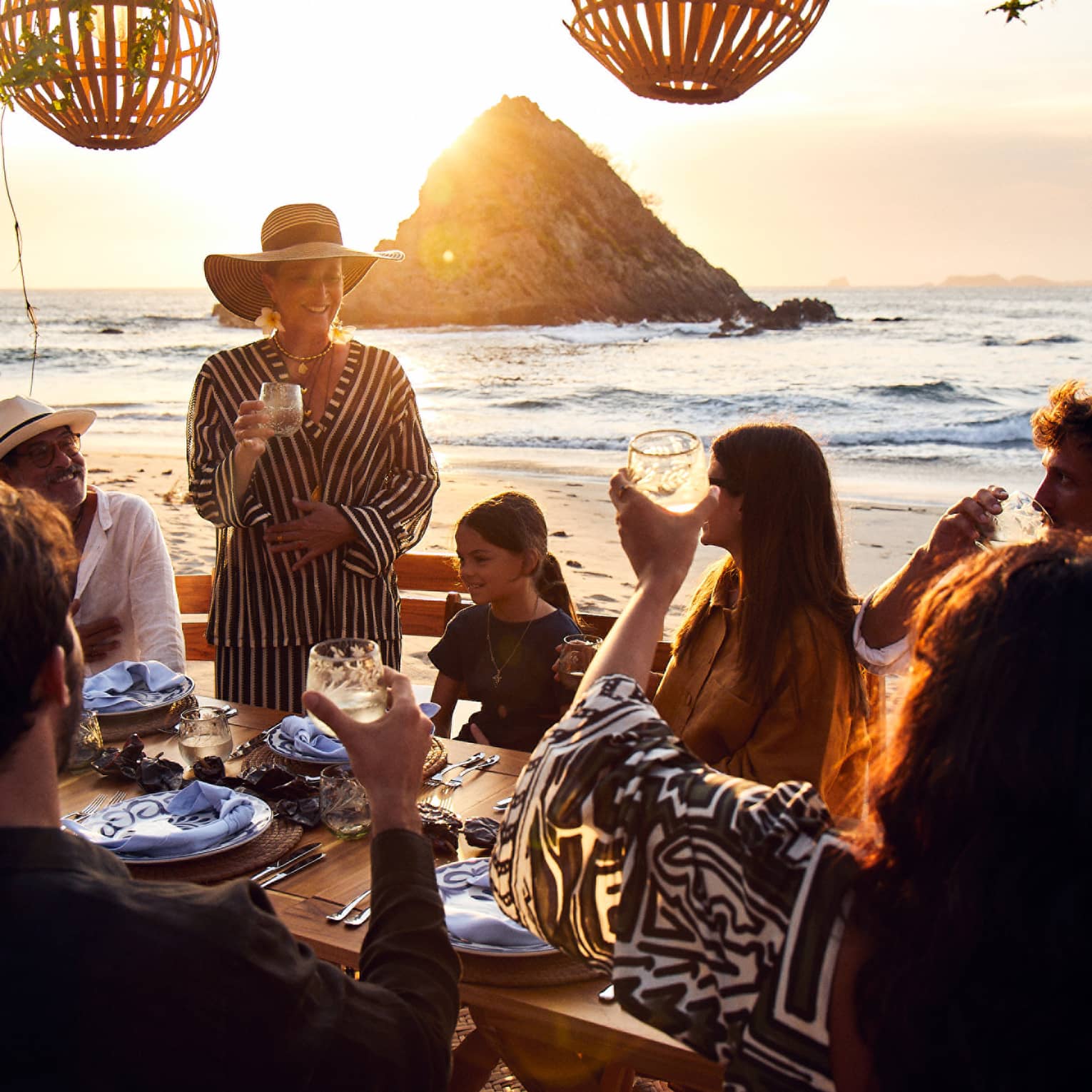 At a set table on the beach, the sun setting over the water behind them, a group of guests raise their glasses in a toast.