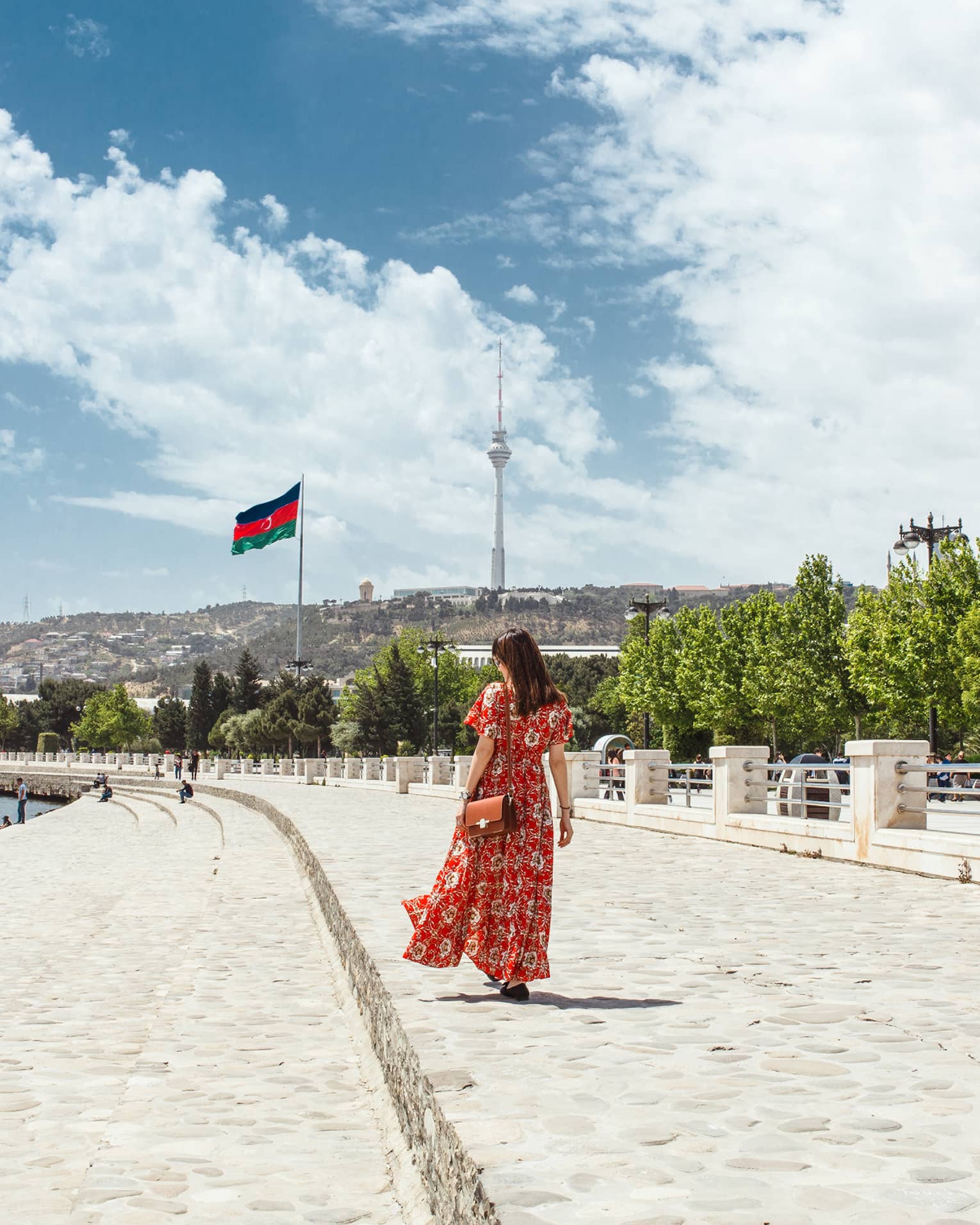 Woman wearing red dress walks along stone waterfront, skyscrapers in background