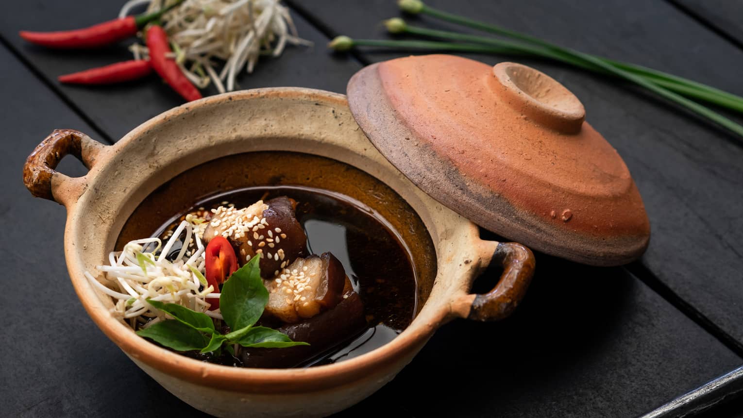 Vietnamese dish in clay pot, garnished with bean shoots, chili, sesame seeds, herbs, with spoon on black surface