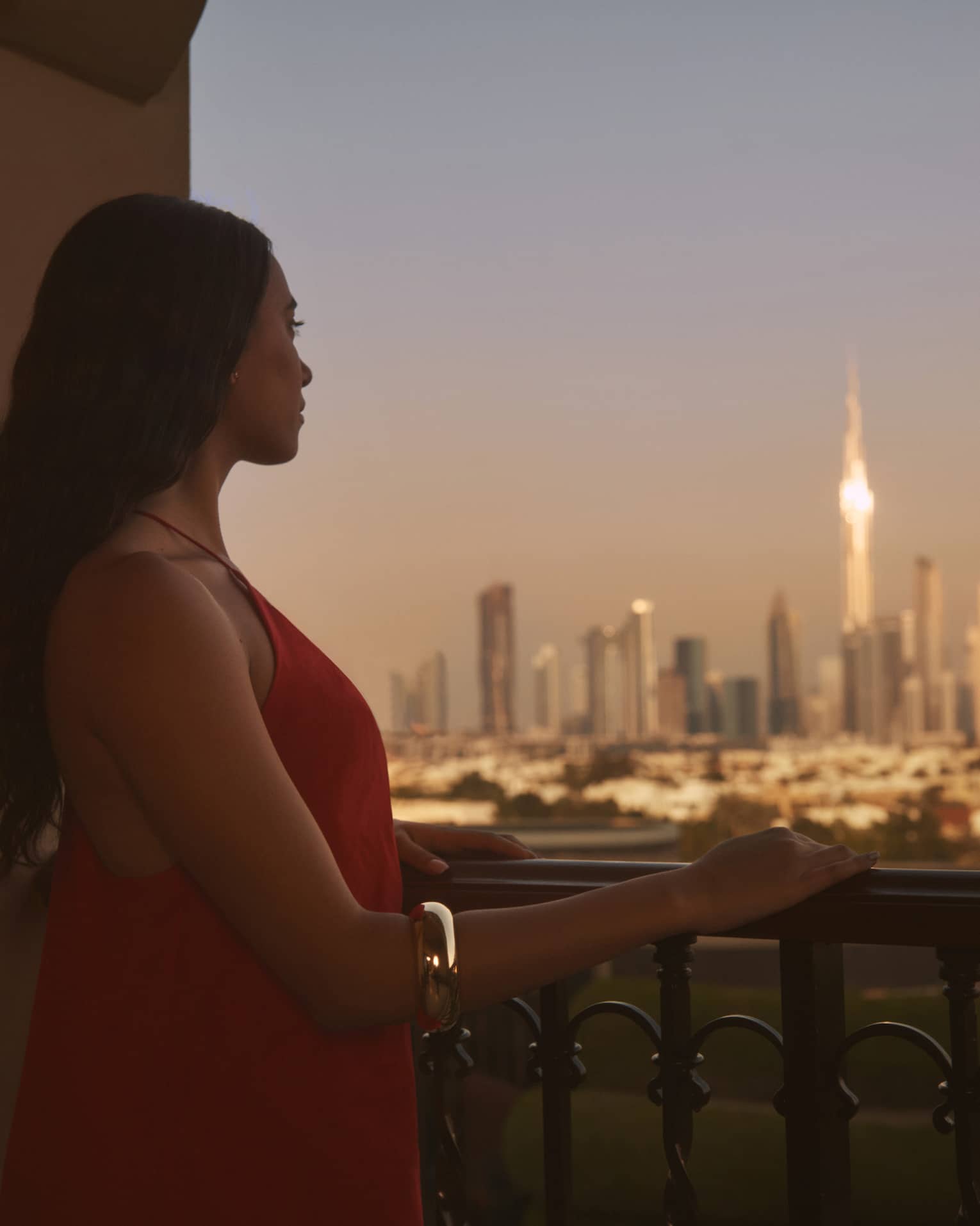Person with long brown hair standing on balcony with hand resting on railing, looking out to a city at dusk