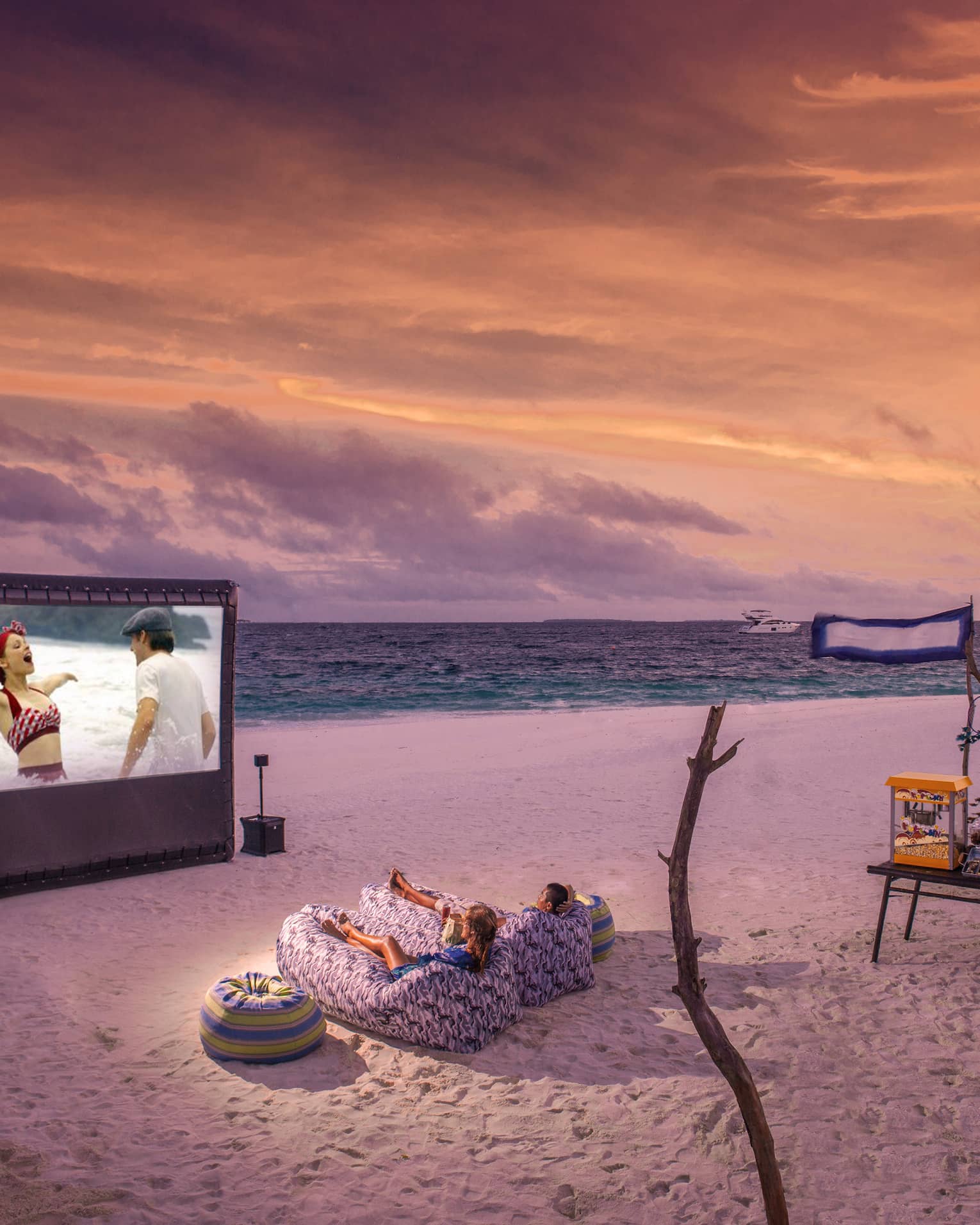 ,Two people in beanbag chairs enjoy a movie night on the beach at dusk, popcorn stand on sand behind them