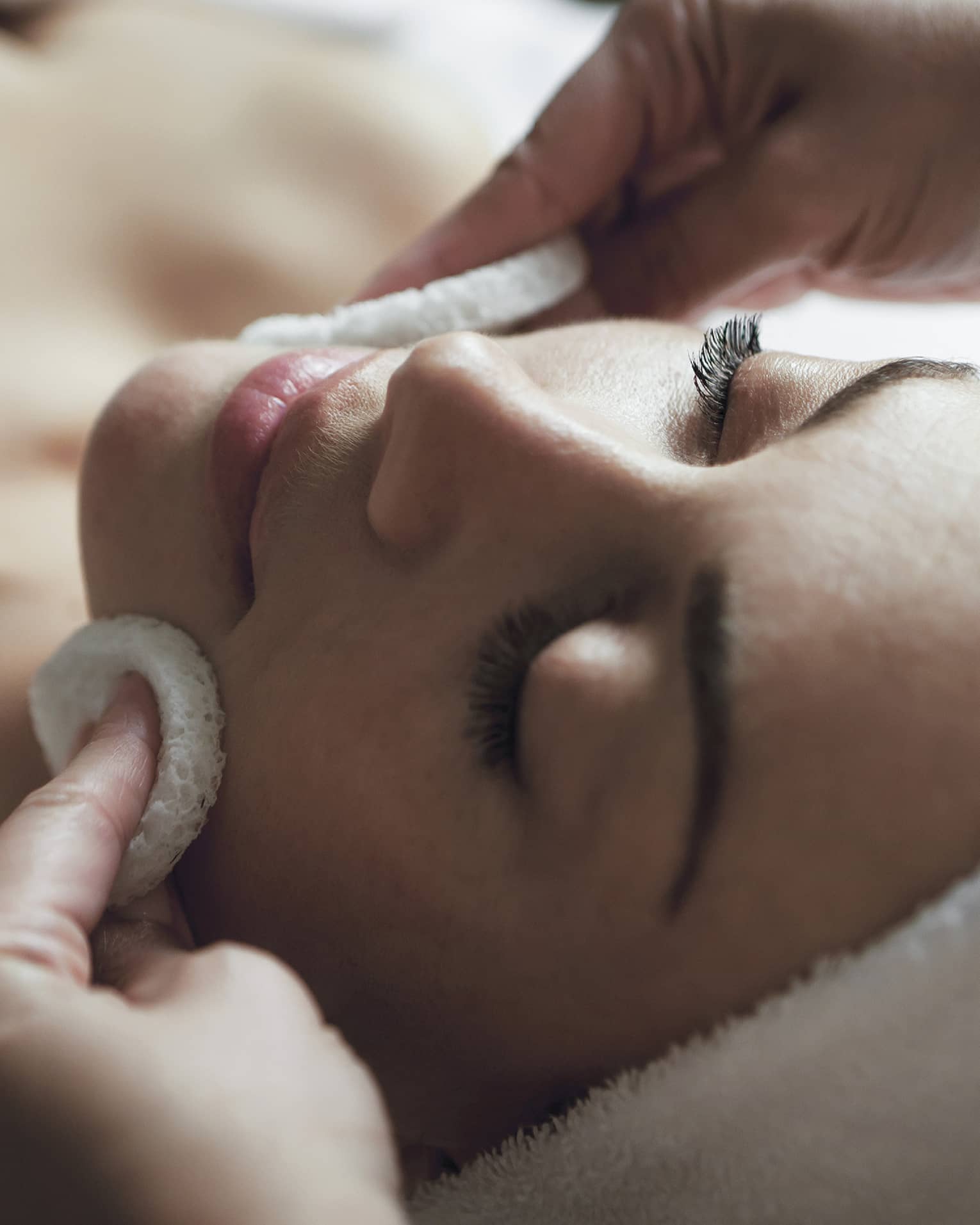 A detail of hands rubbing a sponge on a woman's face as she lies on a massage table with her eyes closed and hair wrapped up in a white towel