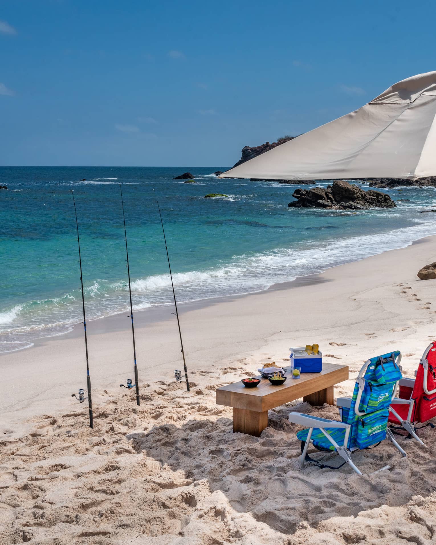 Under an umbrella, drinks, snacks and three colourful beach chairs. Three fishing rods planted in the sand, blue sea ahead.